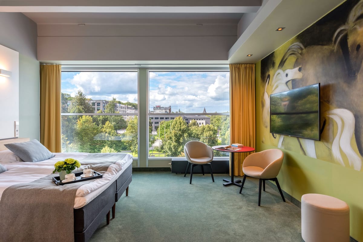 Ein Hotelzimmer mit großem Fenster, einem Bett, einem Schreibtisch, zwei Stühlen und einem Fernseher an der Wand.