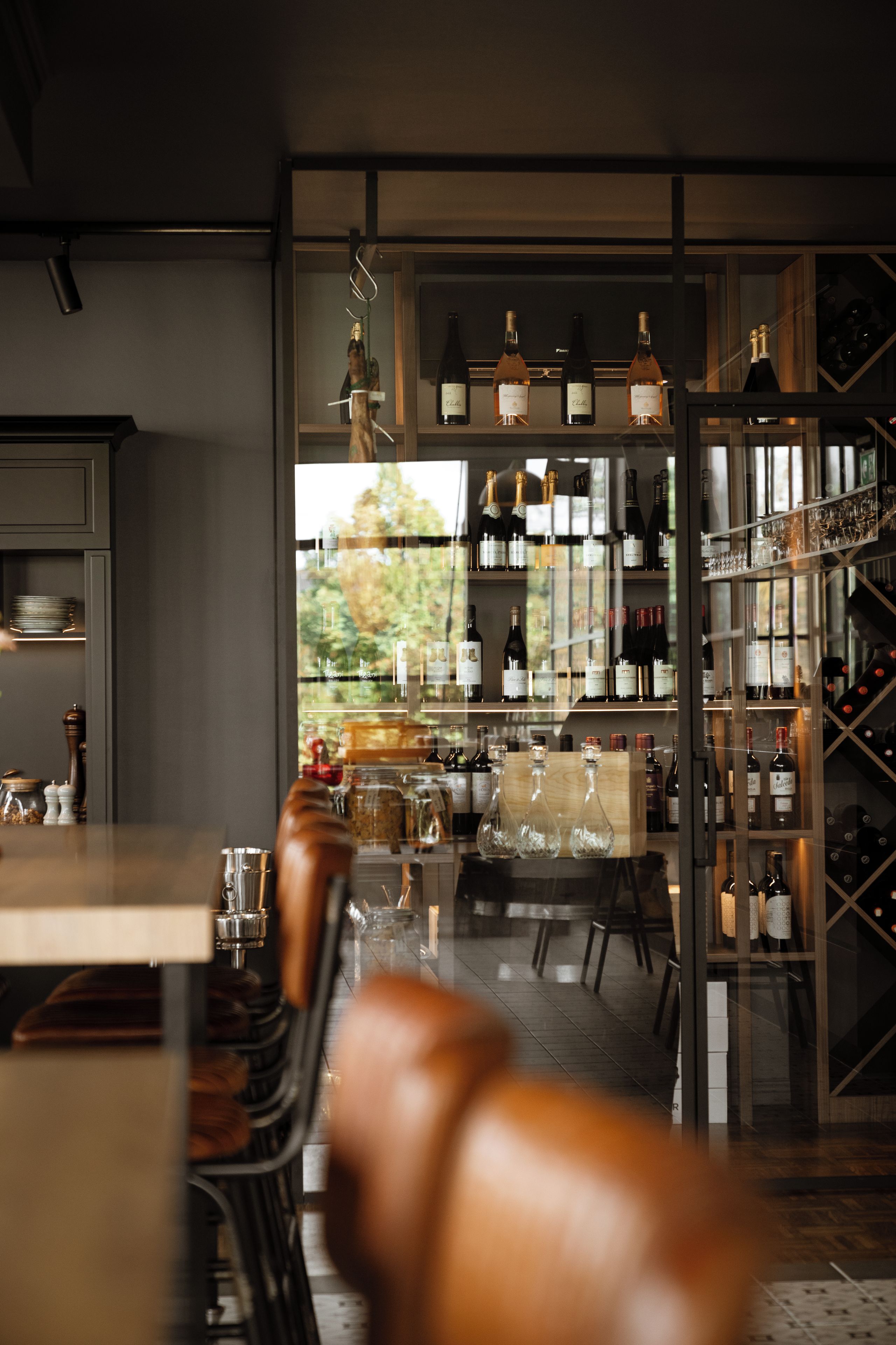 A modern bar area with a variety of wine bottles and glasses on shelves, a wooden table, and a view of the outdoors.