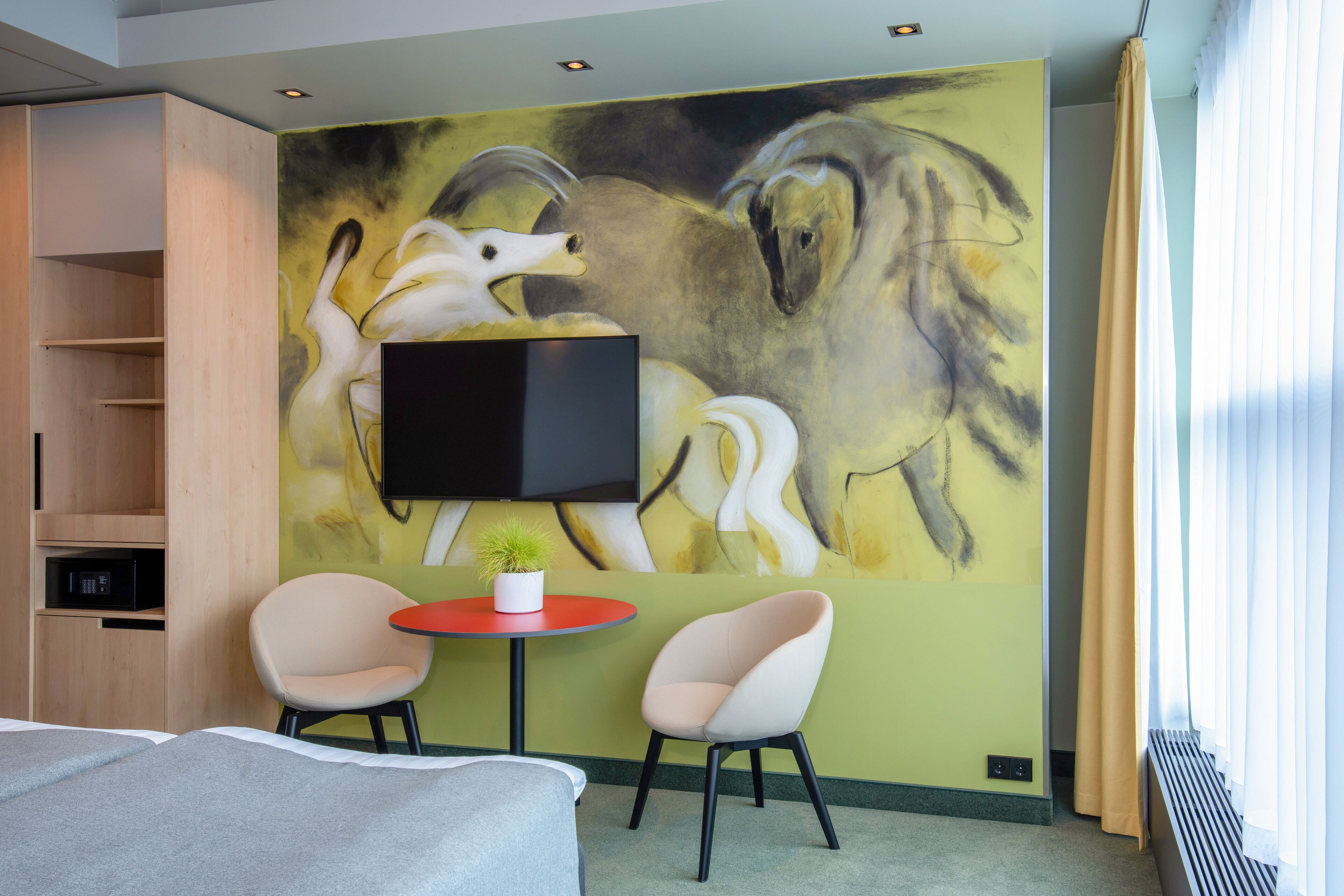 Una habitación de hotel con un cuadro abstracto grande en la pared, un televisor de pantalla plana sobre una pequeña mesa con dos sillas, y una cama con una manta gris.