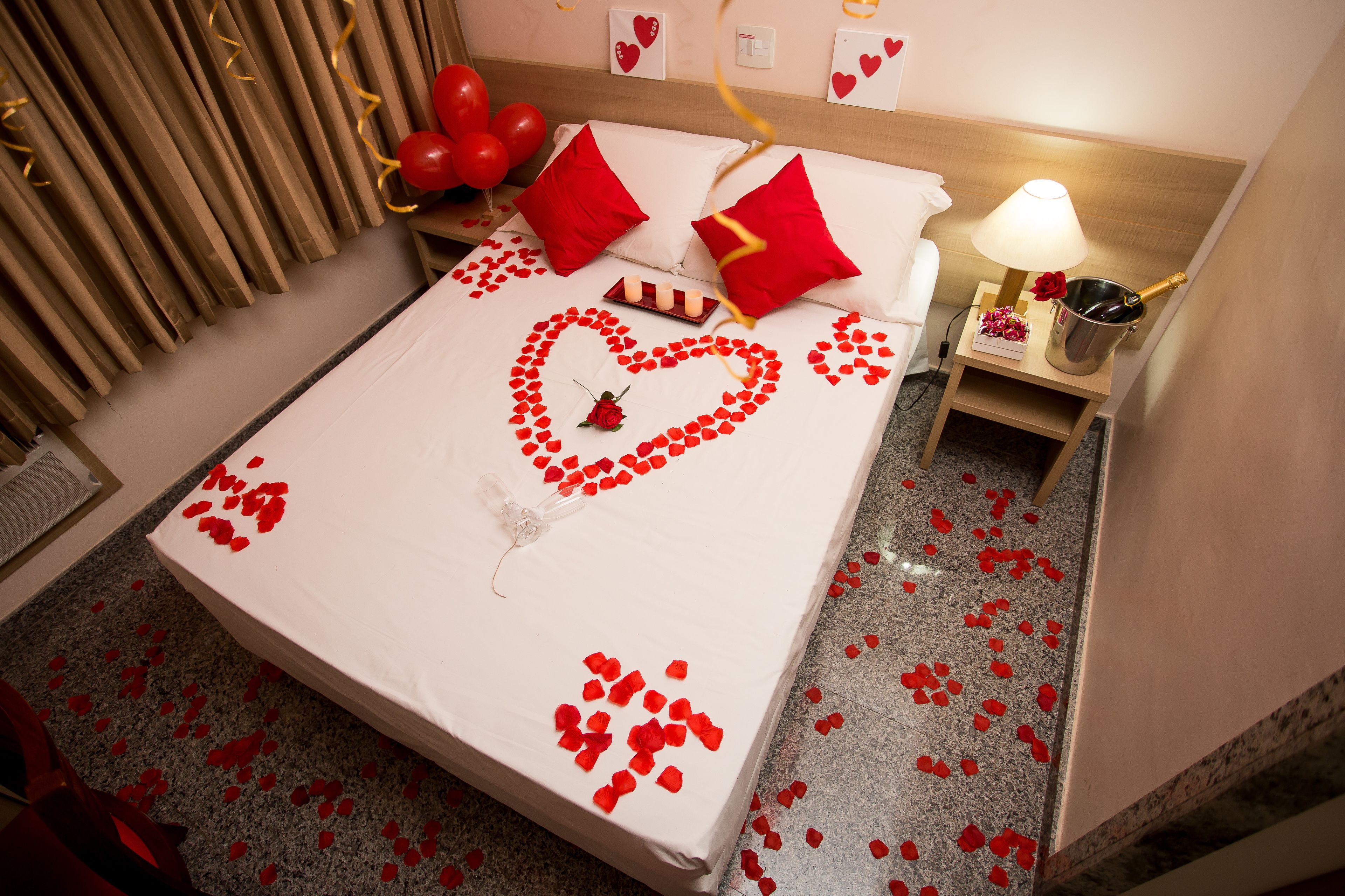 A bed with red heart-shaped decorations, a bedside table with a lamp and a bottle, and a wall with heart decorations.