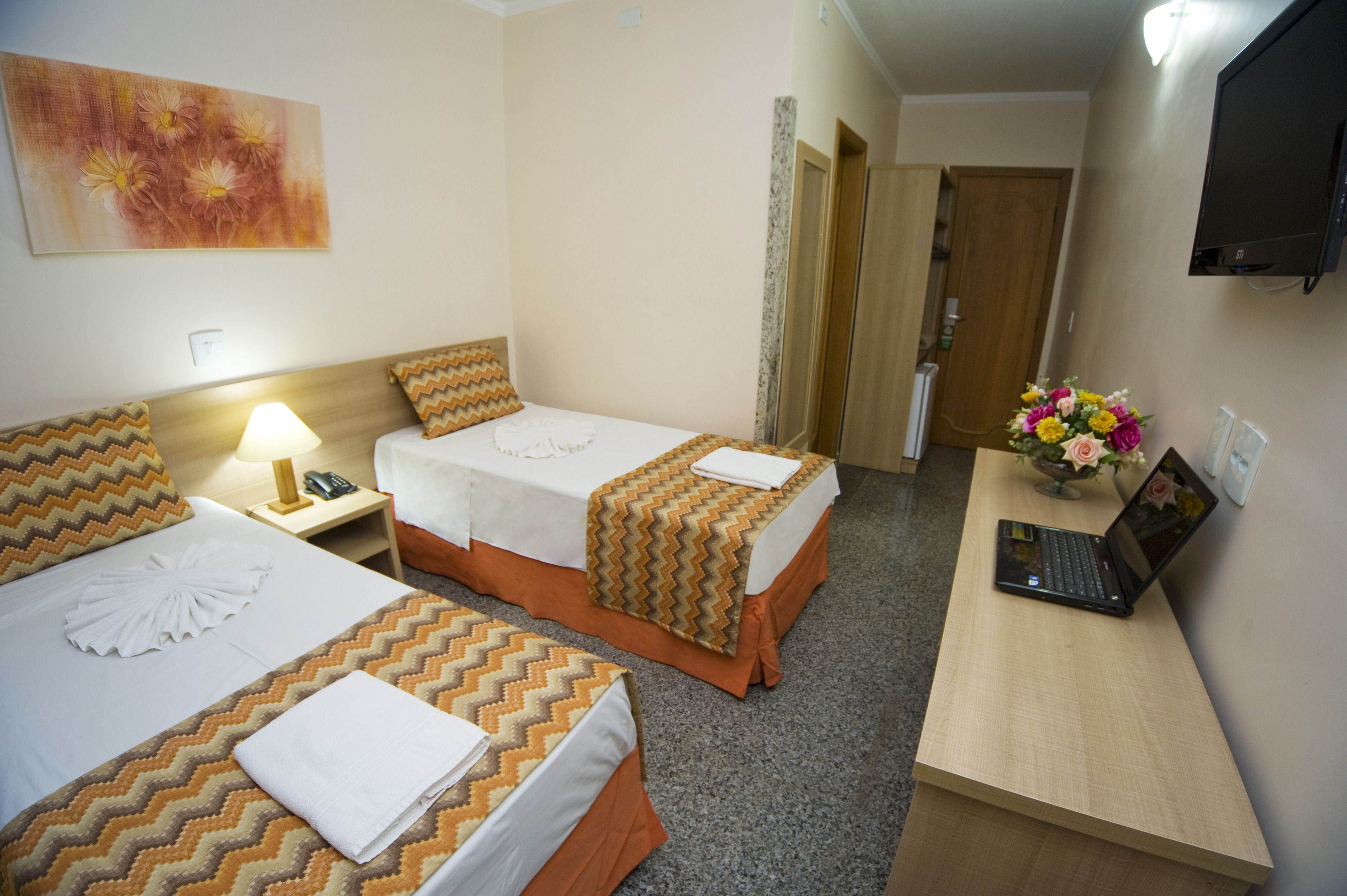 A hotel room with two beds, a desk with a laptop, a TV, and a flower vase.