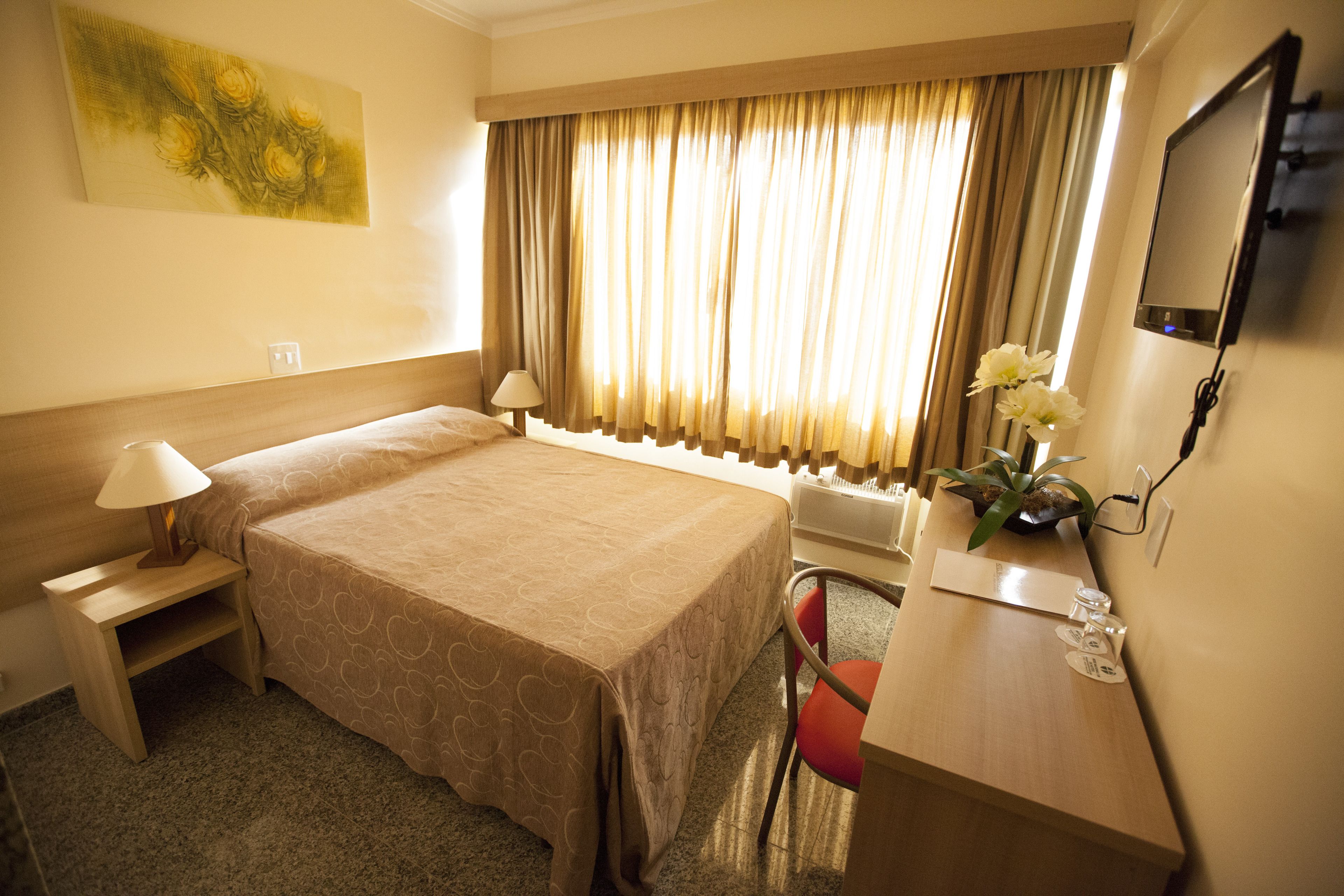 Quarto de hotel com uma cama grande, uma mesa de trabalho com cadeira, televisão e janela com cortinas.