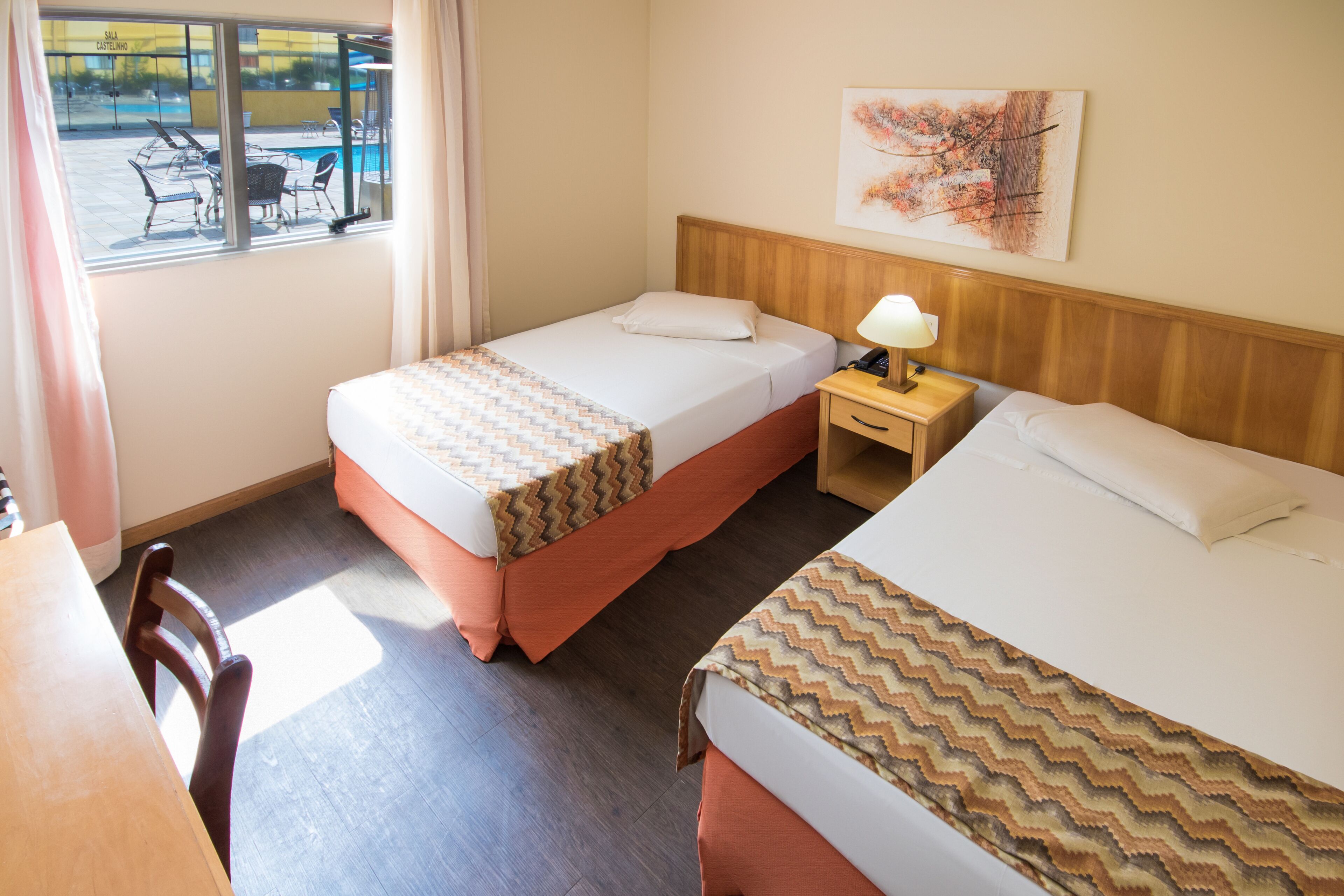 Une chambre d’hôtel avec deux lits, une tête de lit en bois, une table de chevet avec une lampe, une fenêtre donnant sur une piscine extérieure et des chaises, et un tableau au mur.
