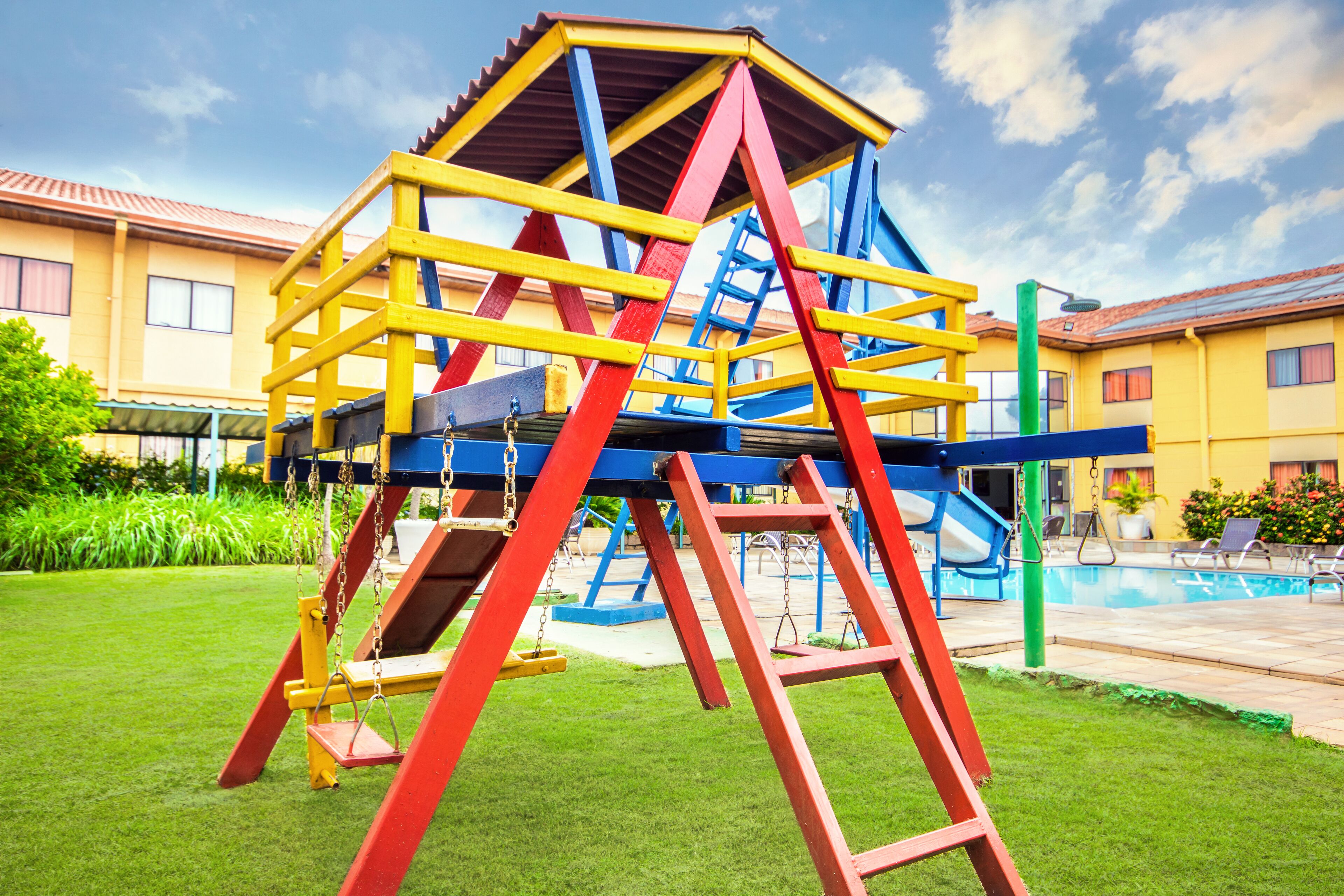 Una estructura de joc colorida amb tobogan i escales d'ascensió, amb piscina i un edifici de sostre enrajolat al fons.