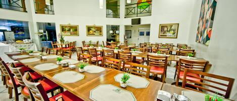 A spacious dining area with multiple tables and chairs, large windows, and artwork on the walls.