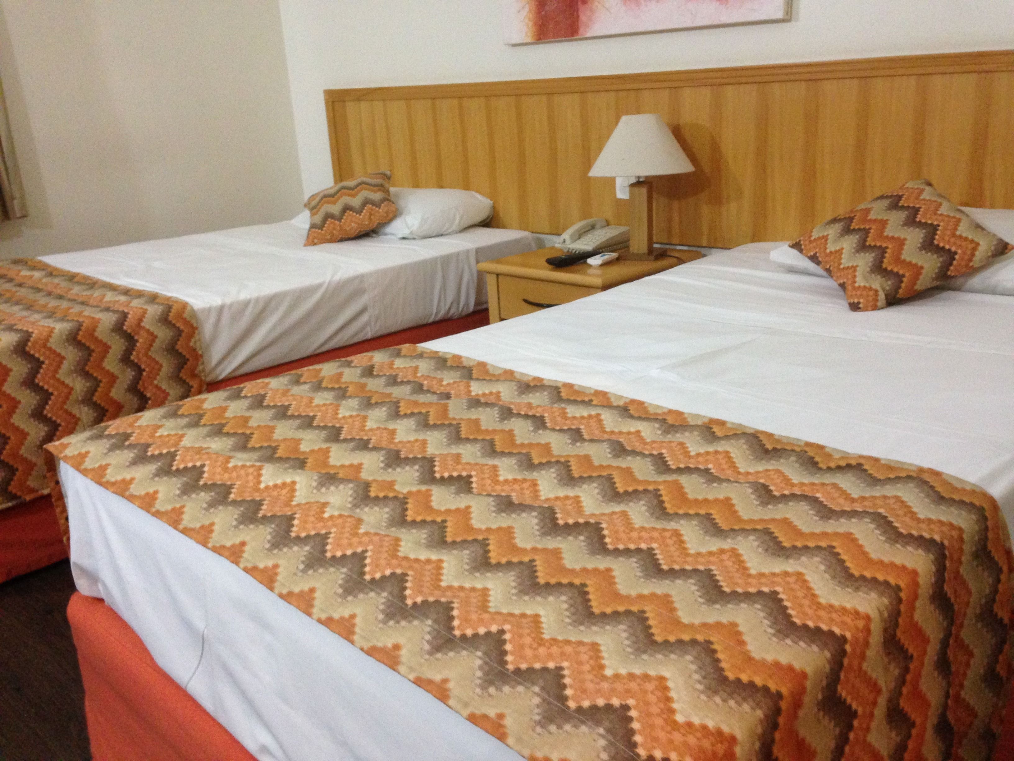 Een hotelkamer met twee bedden, elk voorzien van een beddeken en kussens. Er staat een nachtkastje met een lamp en een telefoon.