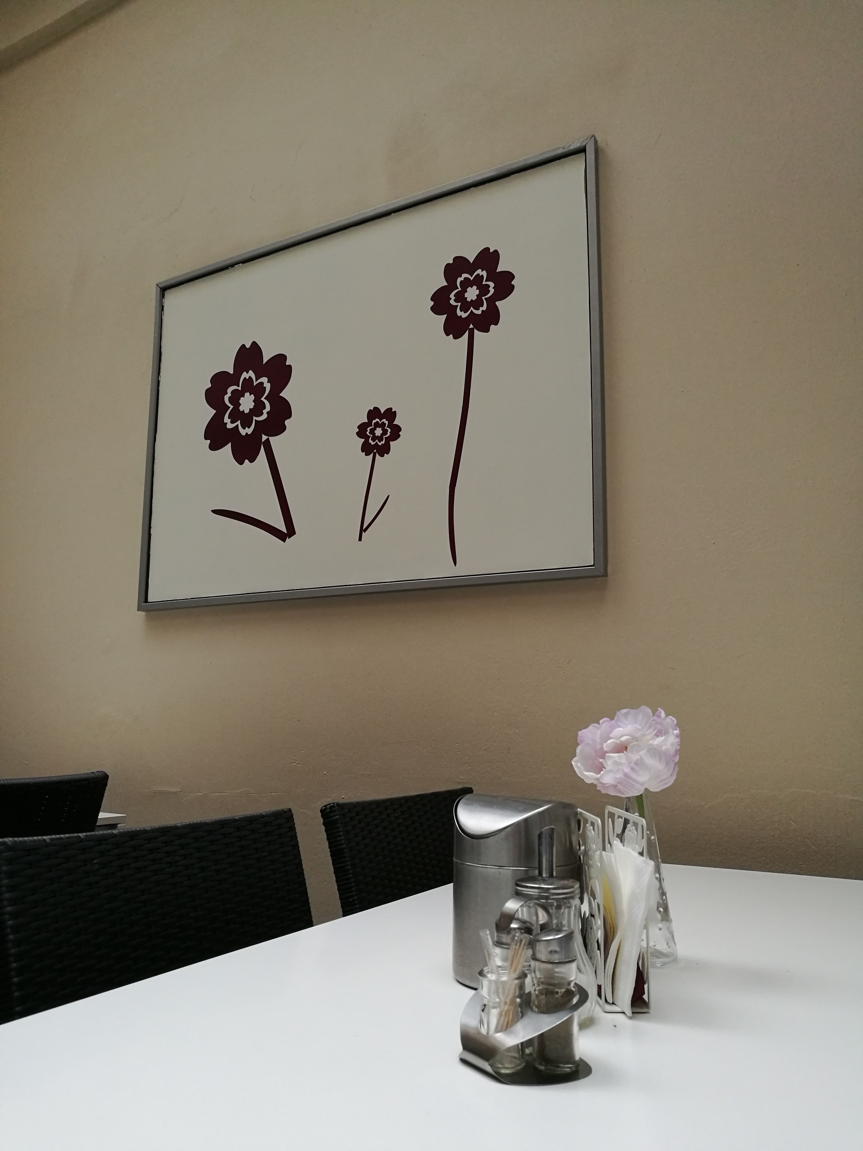 Una mesa de comedor con una obra de arte floral enmarco, un jarro metálico y un florero con una flor.