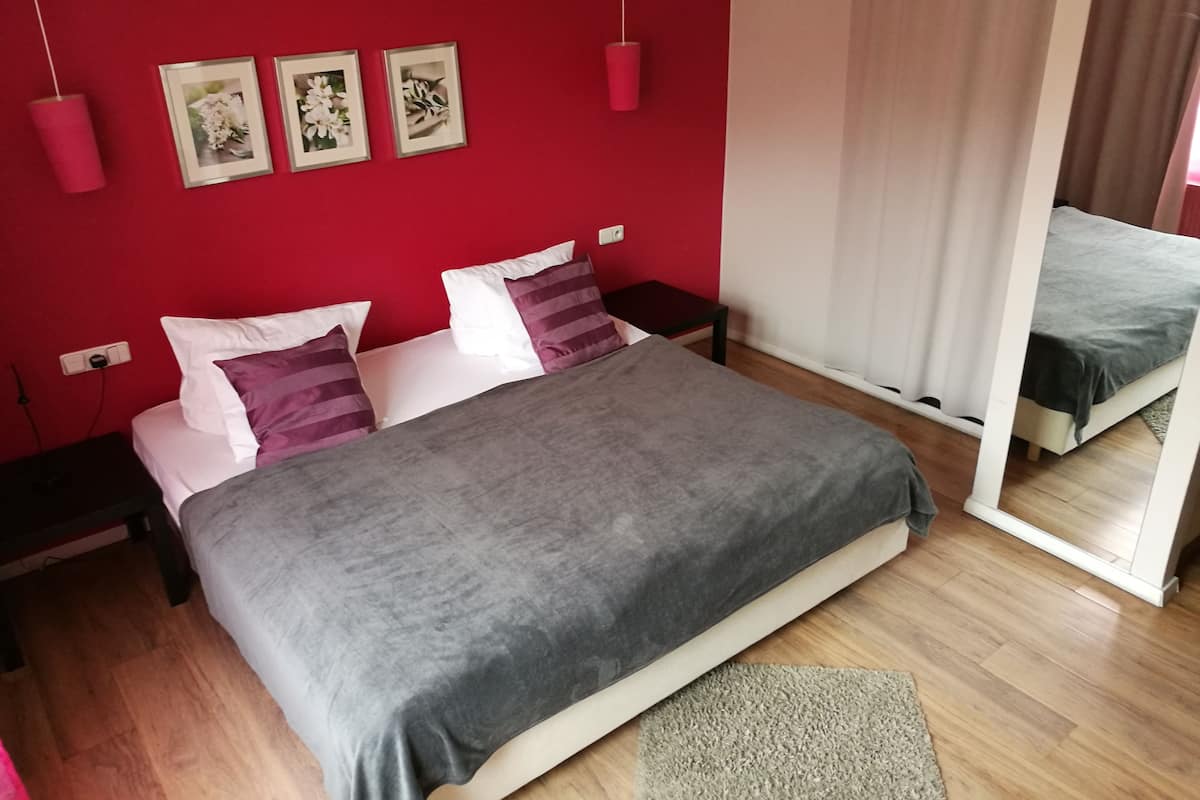 Quarto com parede vermelha, cama com cobertor cinza, almofadas brancas e roxas, uma mesinha de cabeceira pequena e um espelho refletindo a cama.