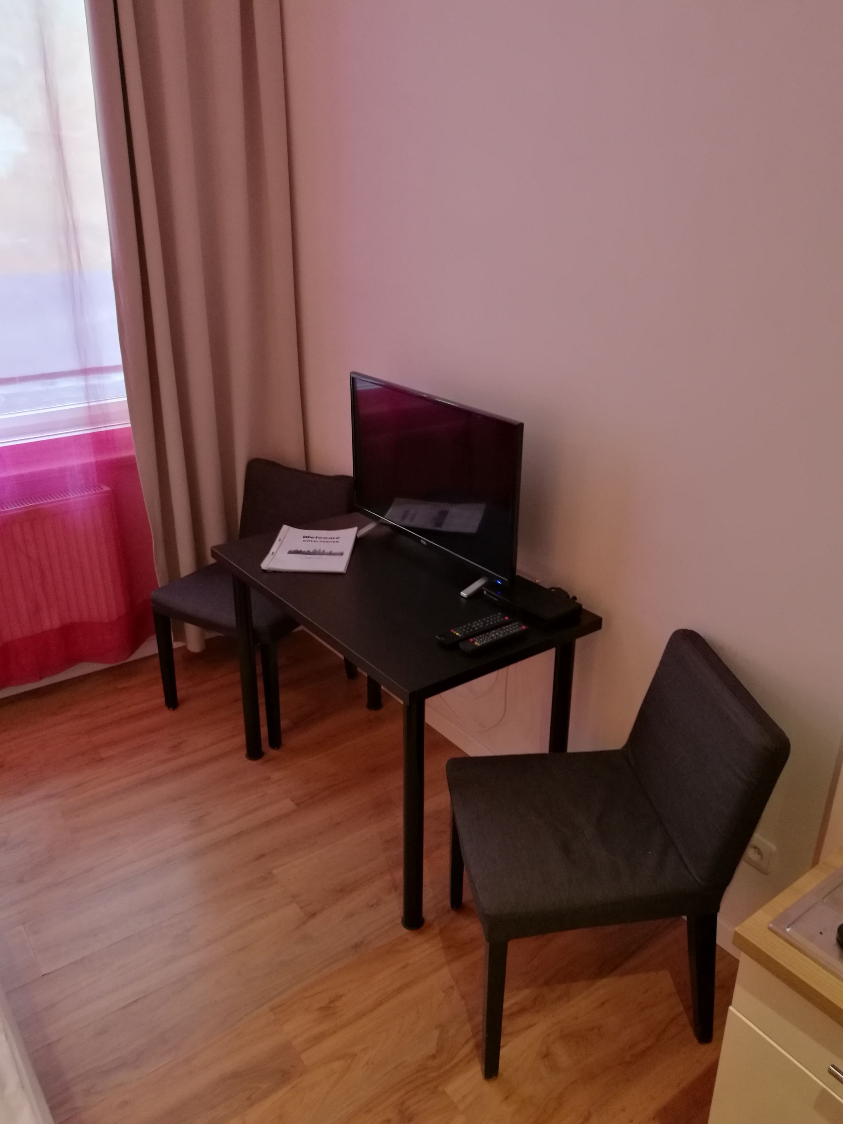 Una habitación pequeña con escritorio, una silla, televisión y ventana con cortinas.