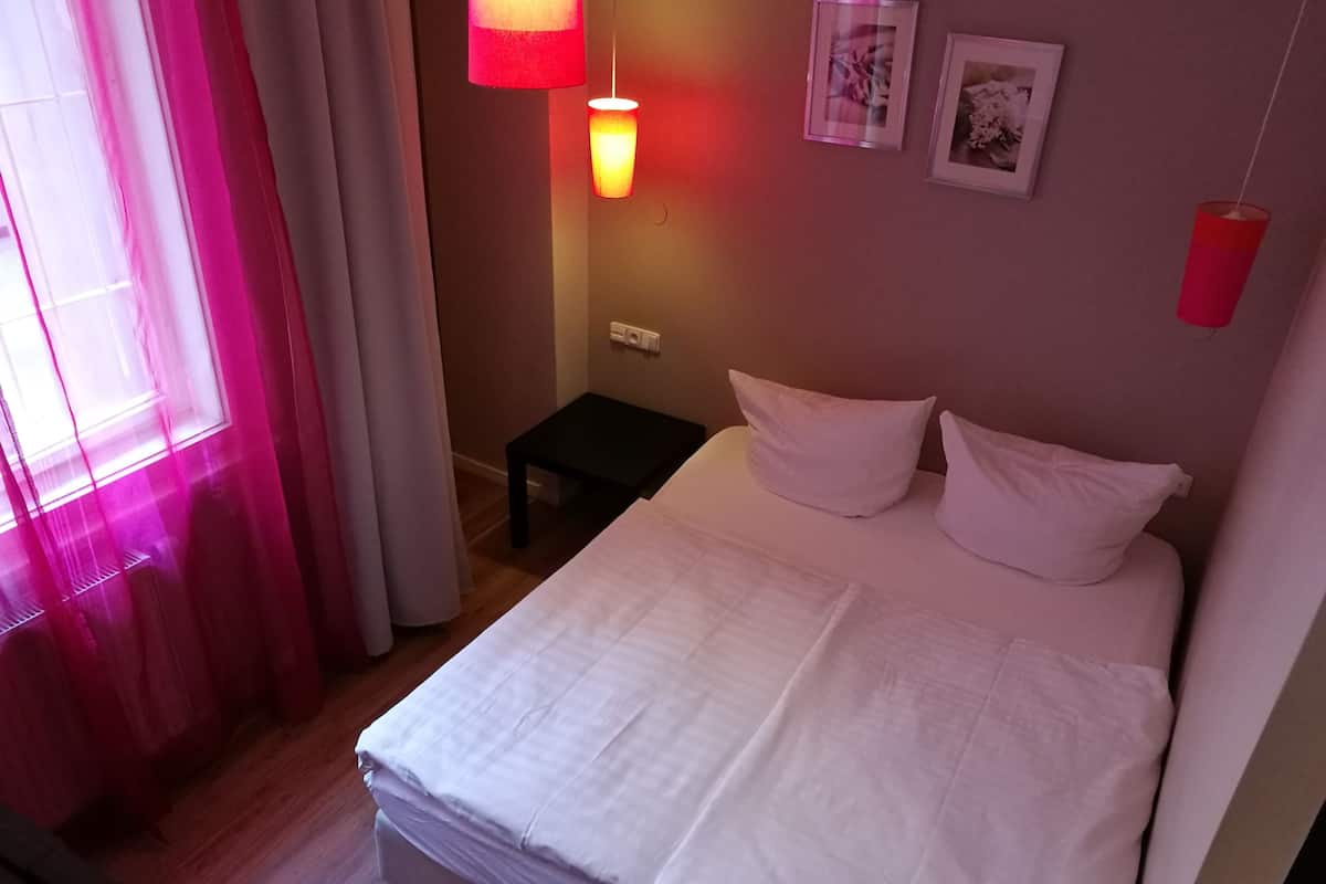 Cama bem arrumada com roupa de cama branca, uma pequena mesa de cabeceira e duas luminárias fixadas na parede.