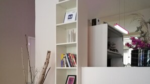 Uma sala de estar moderna com um sofá preto, uma almofada vermelha, uma estante branca e um vaso com flores.