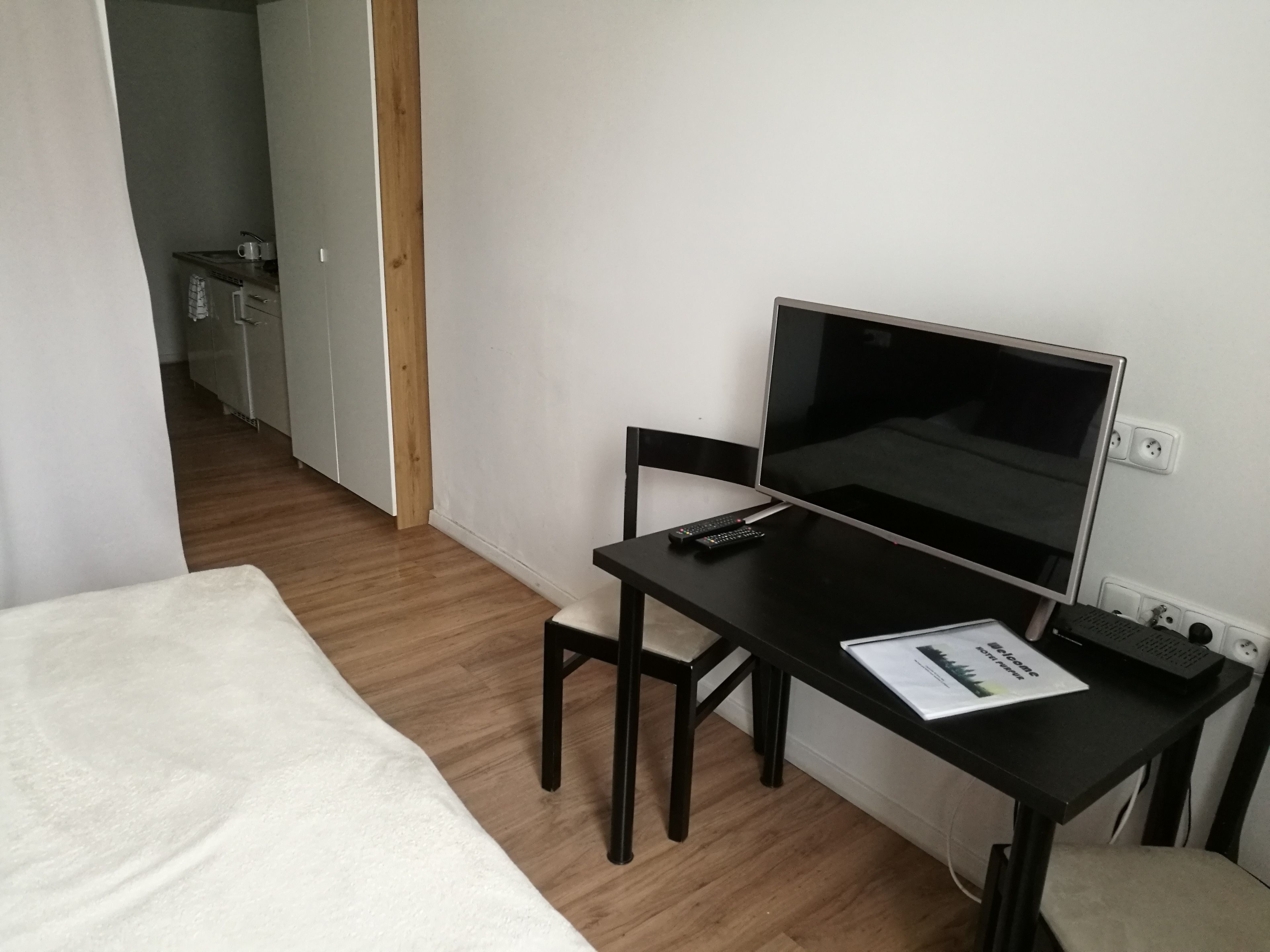 Ein kleines, ordentliches Zimmer mit einem Bett, einem Schreibtisch mit Computer und einem Stuhl. Auf dem Schreibtisch liegt eine Fernbedienung und auf dem Tisch eine Broschüre.