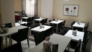 Uma área de refeições com mesas brancas e cadeiras pretas, uma janela grande com cortinas e um quadro de flores na parede.