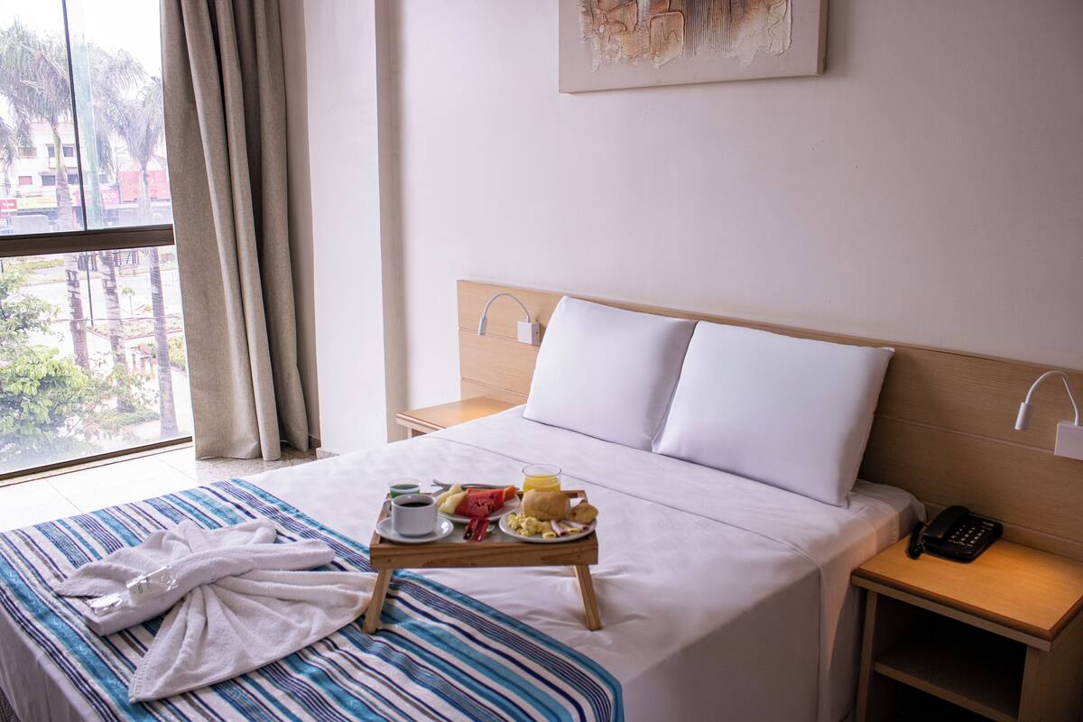 A hotel room with a bed, a bedside table with a phone, a tray with breakfast items, and a window with curtains.