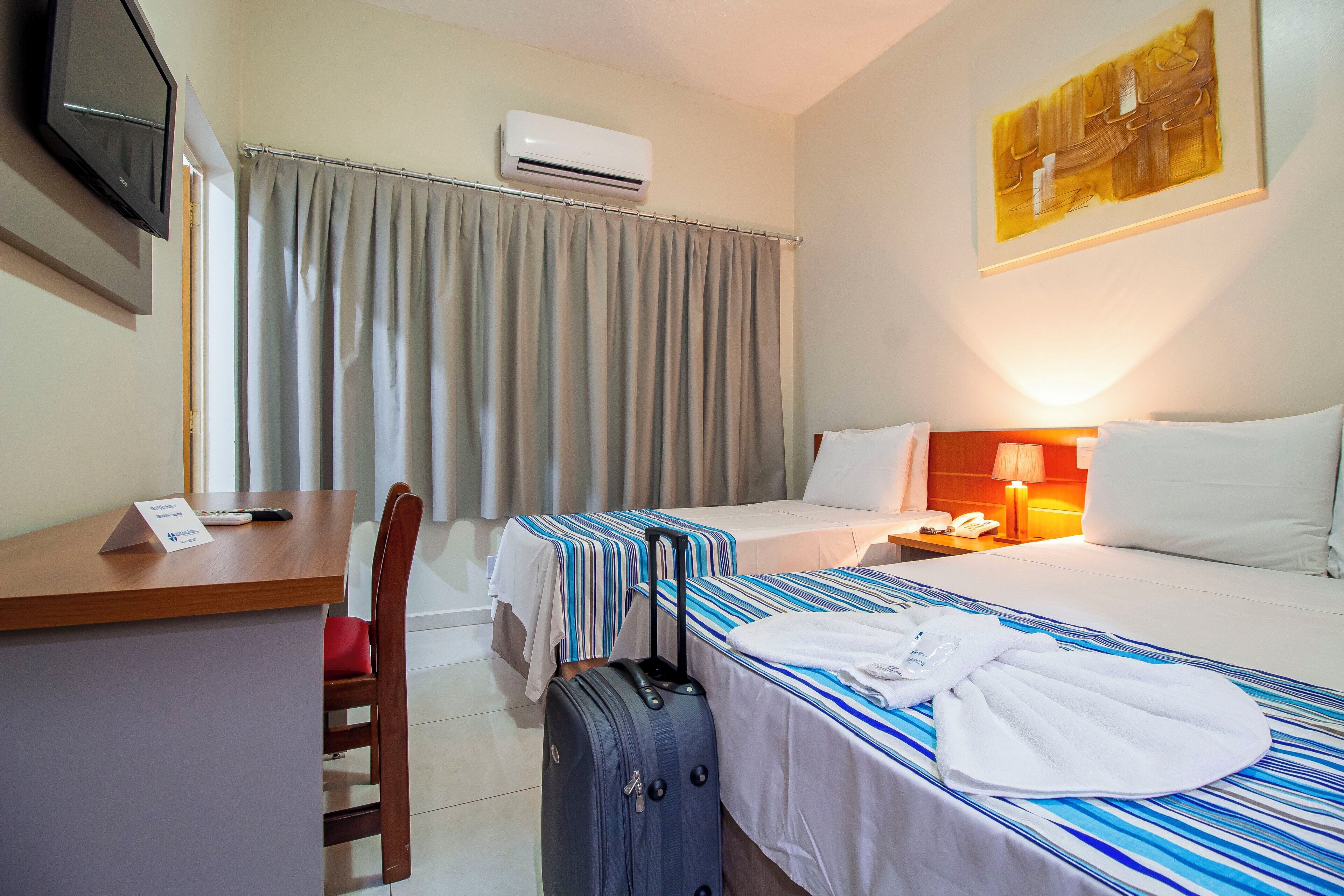 A hotel room with two beds, a desk, a chair, a TV, an air conditioner, and a painting on the wall.