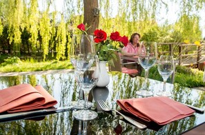 An outdoor dining table with clear glassware, a vase with red flowers, and a person seated in the background.