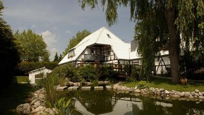 A traditional half-timbered house with a pond in front, surrounded by greenery and trees. - Gartenhotel Weisser Hof (Malente)