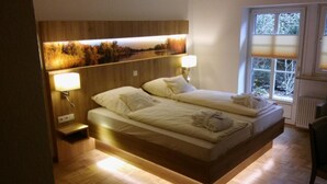 A modern bedroom with a large bed, two bedside lamps, a view of trees through a window, and a wooden headboard. - Gartenhotel Weisser Hof (Malente)