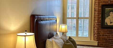 A bedroom with a brick wall, a window with blinds, a bed with patterned bedding, and two bedside lamps.