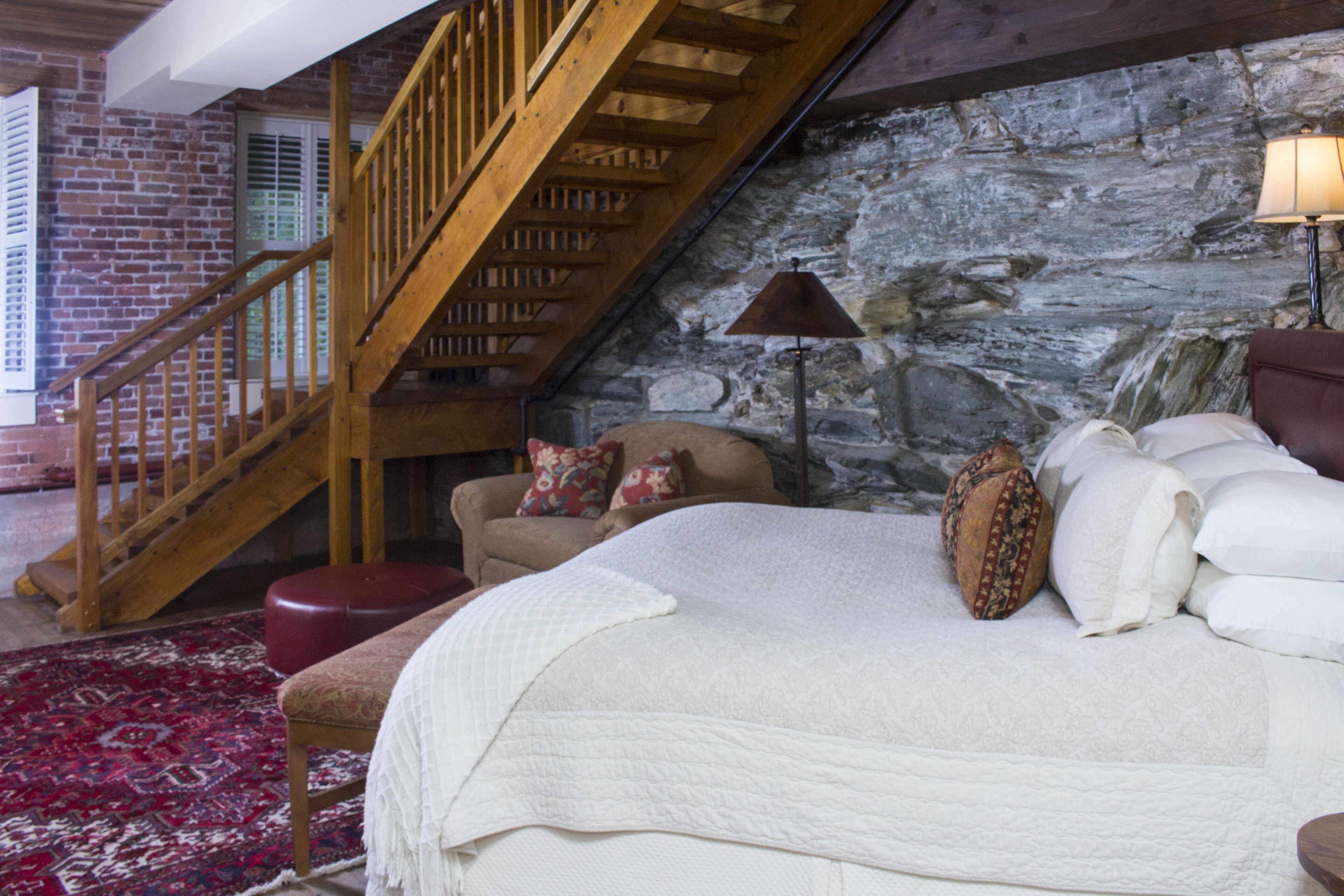 A bedroom with a large bed, a red ottoman, a wooden staircase, and a stone wall.