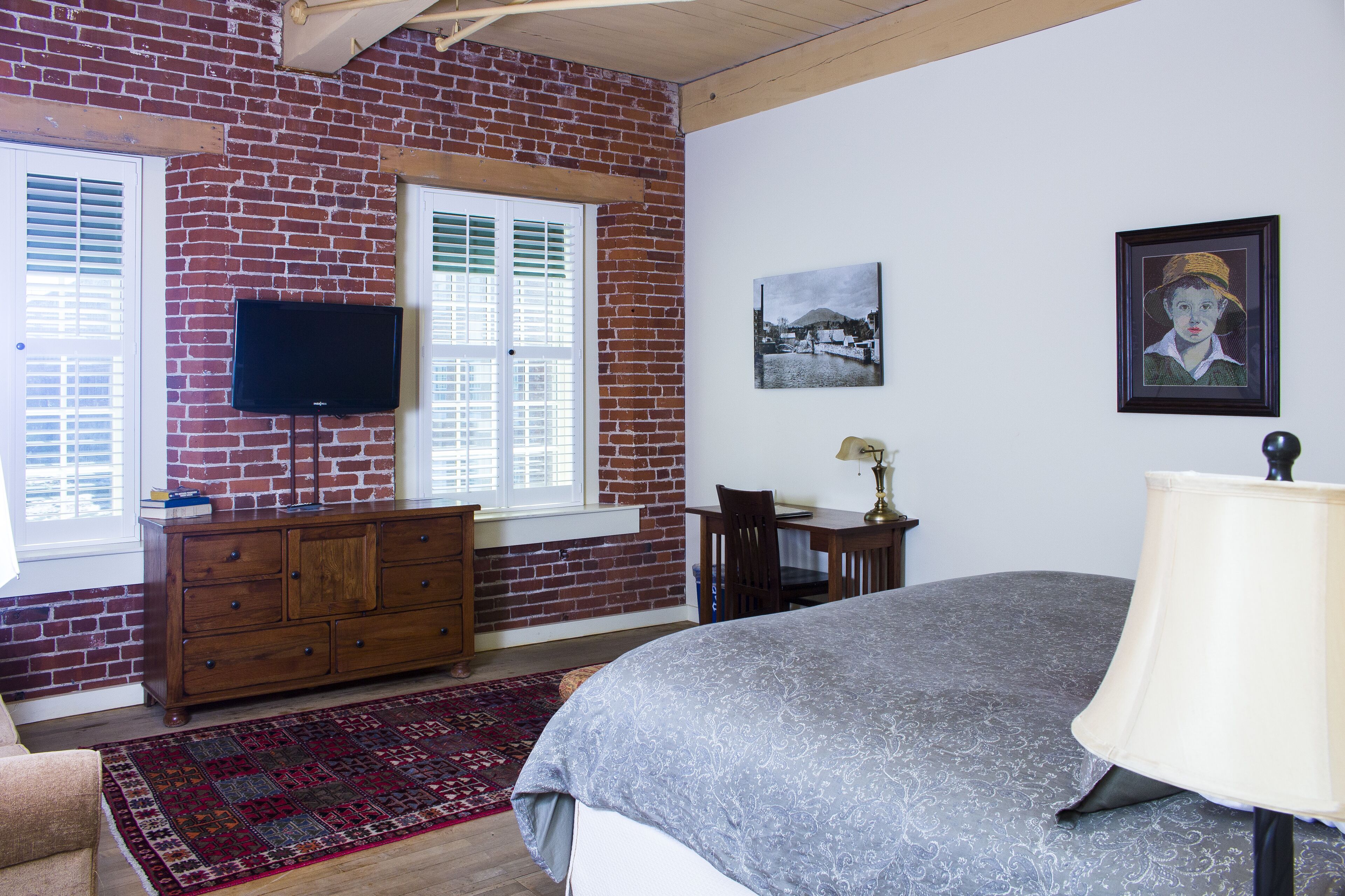 Un dormitorio con pared de ladrillo, cama, escritorio, silla, televisor y obras de arte en la pared.