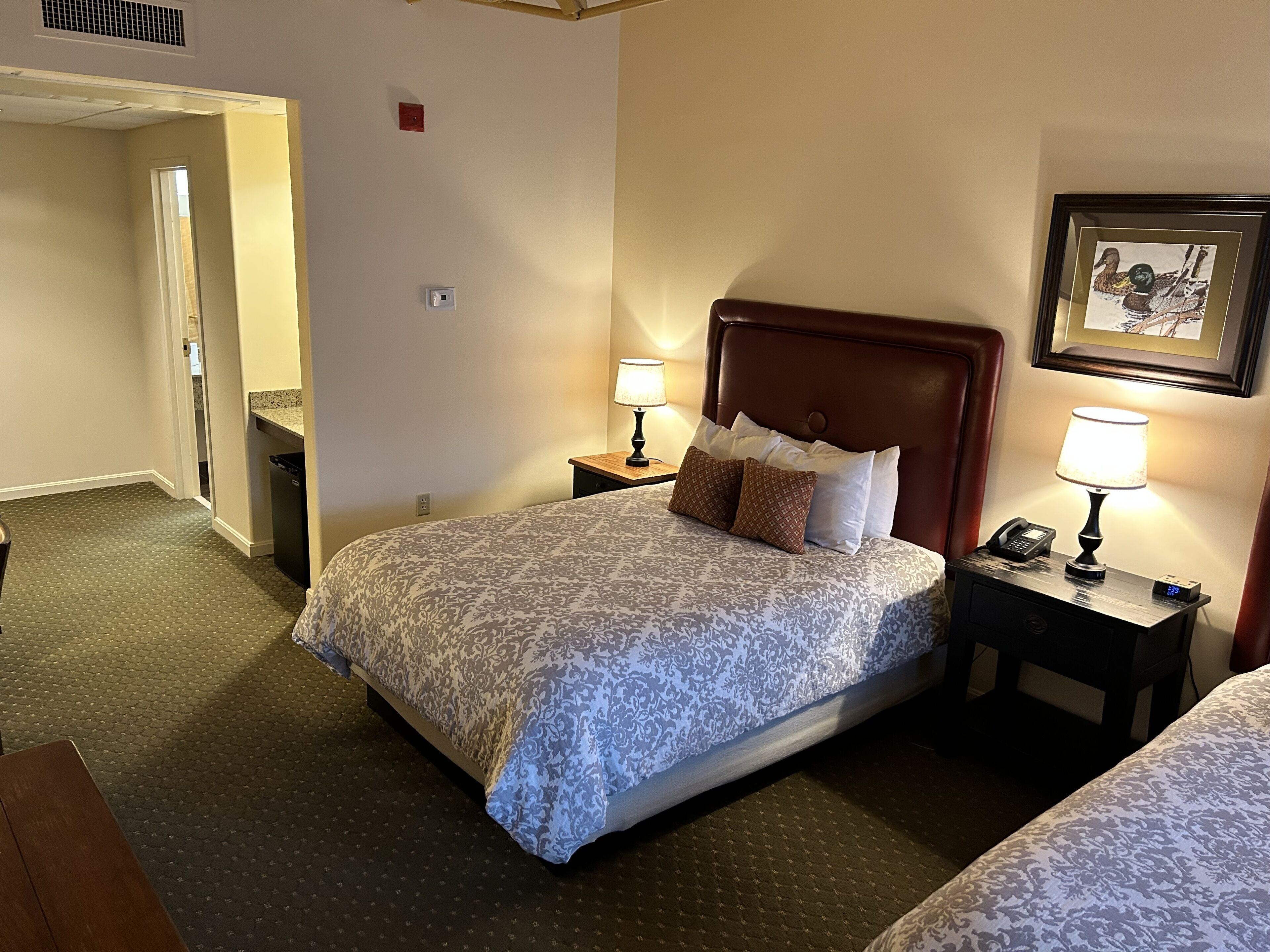 Una habitación de hotel con una cama, dos mesitas de noche con lámparas, un cuadro en la pared y una puerta que da a otra estancia.