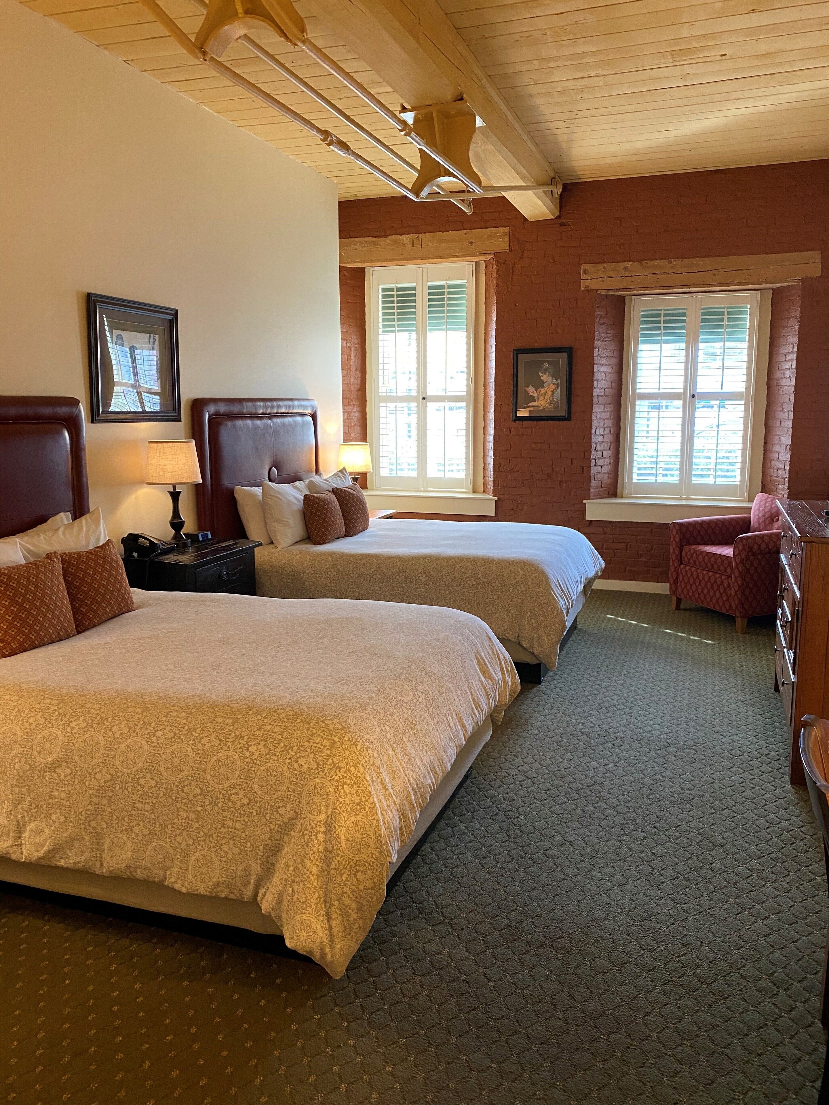 A hotel room with two beds, a desk, and a chair. There are windows with shutters and a ceiling with exposed beams.