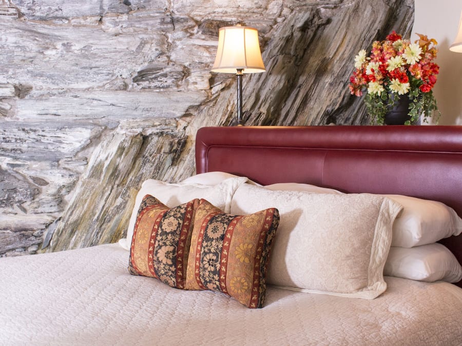 Un dormitorio con pared de piedra, cabecera roja y cama con muchos almohadones.