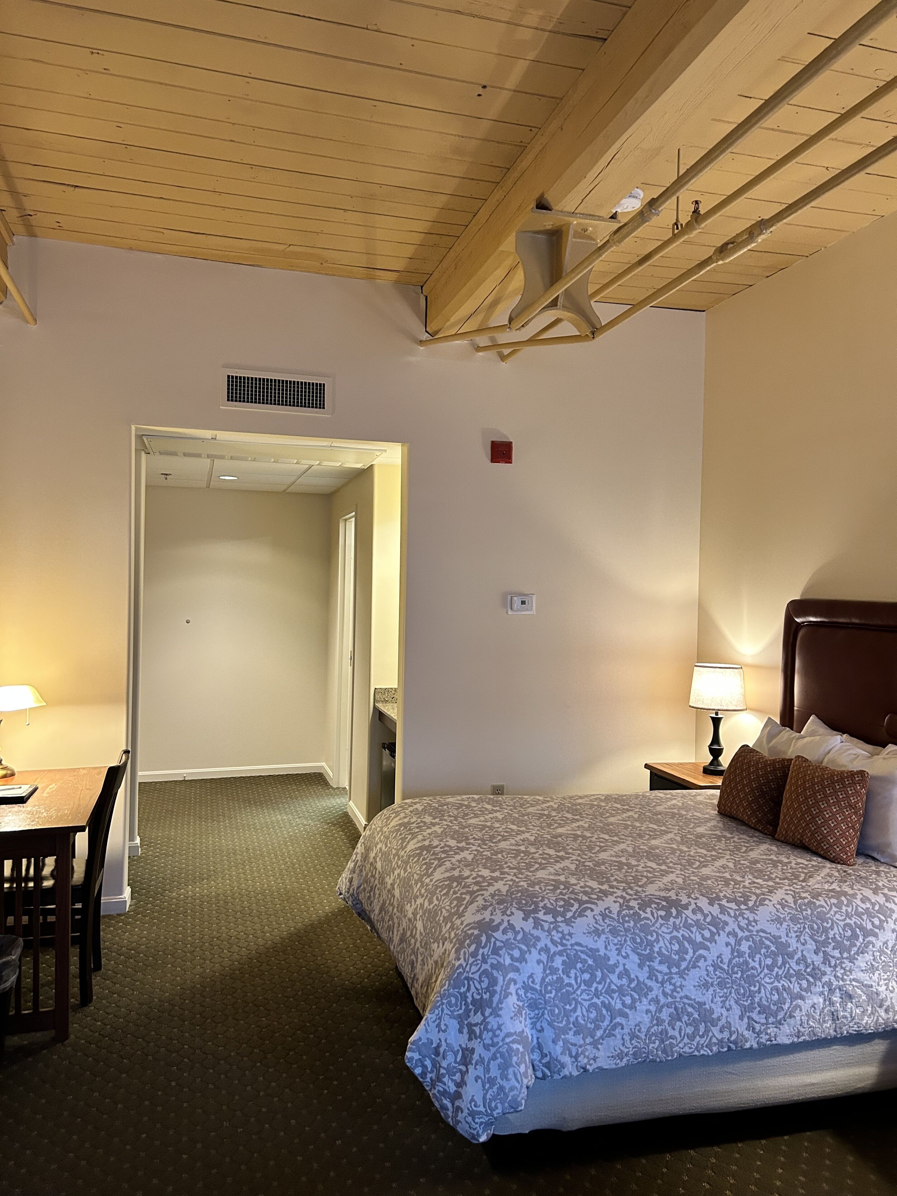 A hotel room with a bed, a desk, and a chair. There is a door leading to another room.