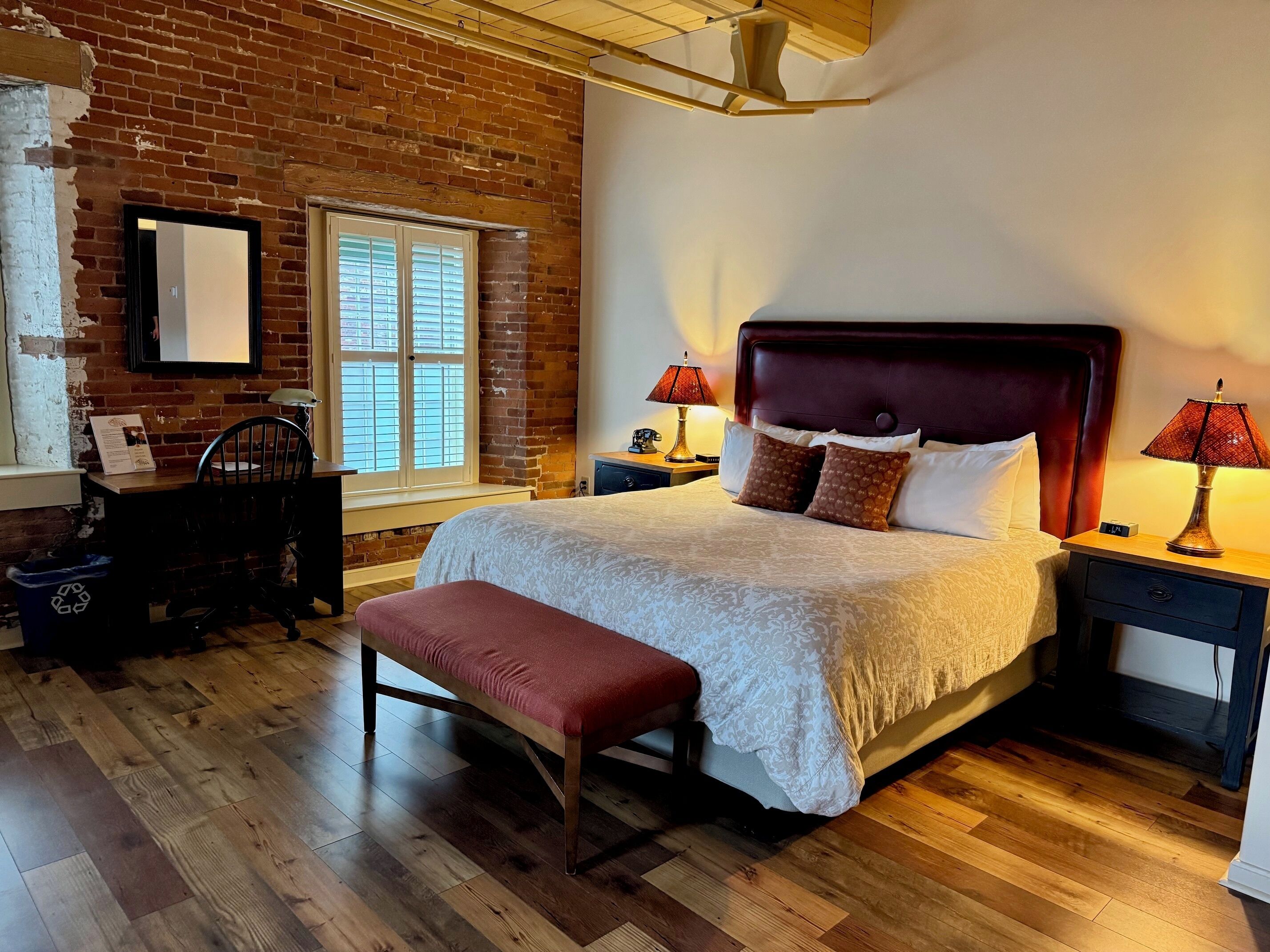 A hotel room with a large bed, a wooden bench, a desk with a chair, a dresser, and a window with blinds.