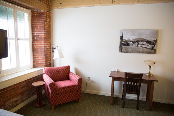 A room with a brick wall, a window with blinds, a pink patterned chair, a wooden desk with a lamp, and a black and white painting on the wall. - The Common Man Inn & Restaurant (Claremont)