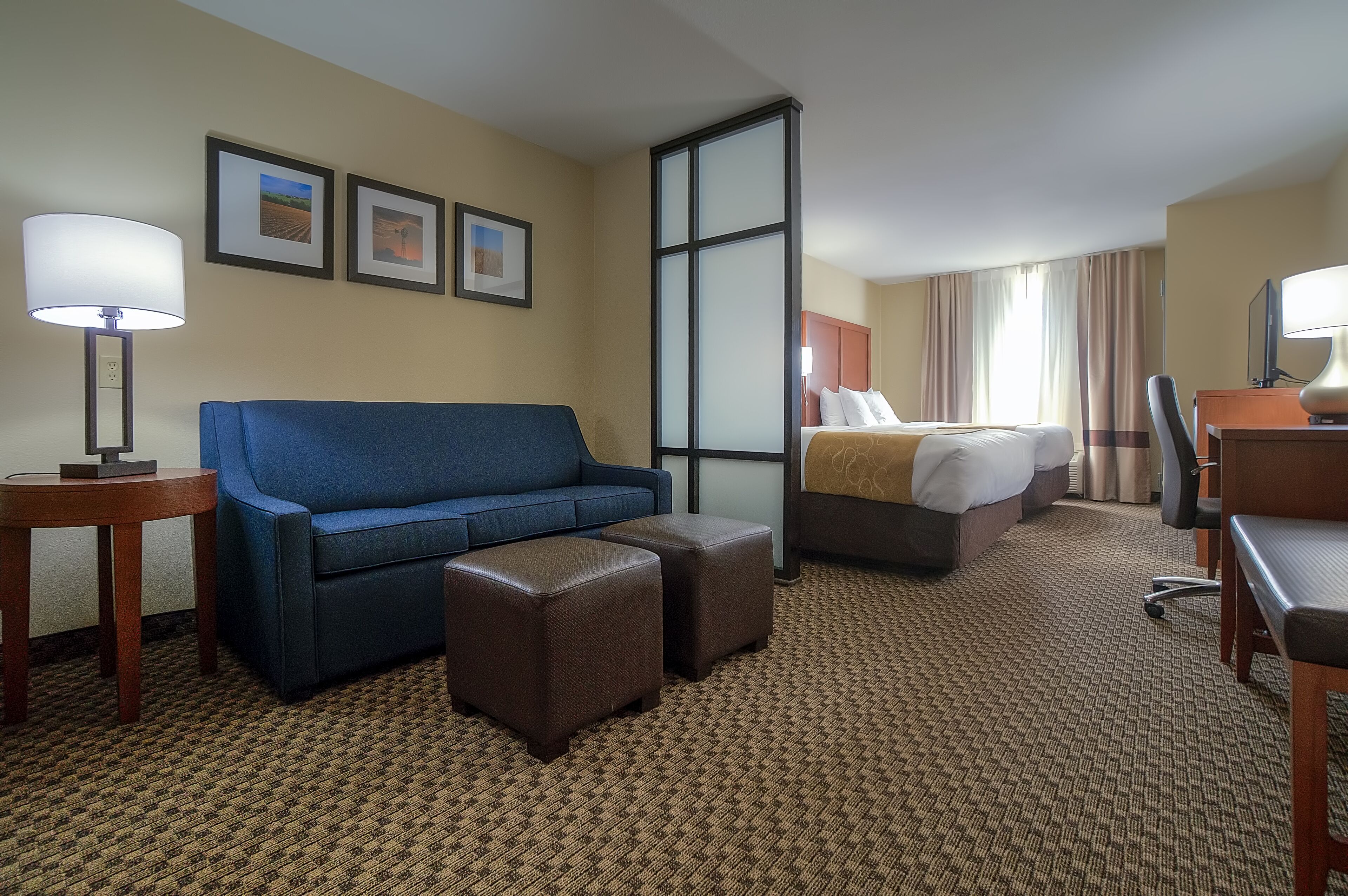 Habitación de hotel con sofá azul, cama con cabecero de madera, escritorio con silla y un ventanal con cortinas.