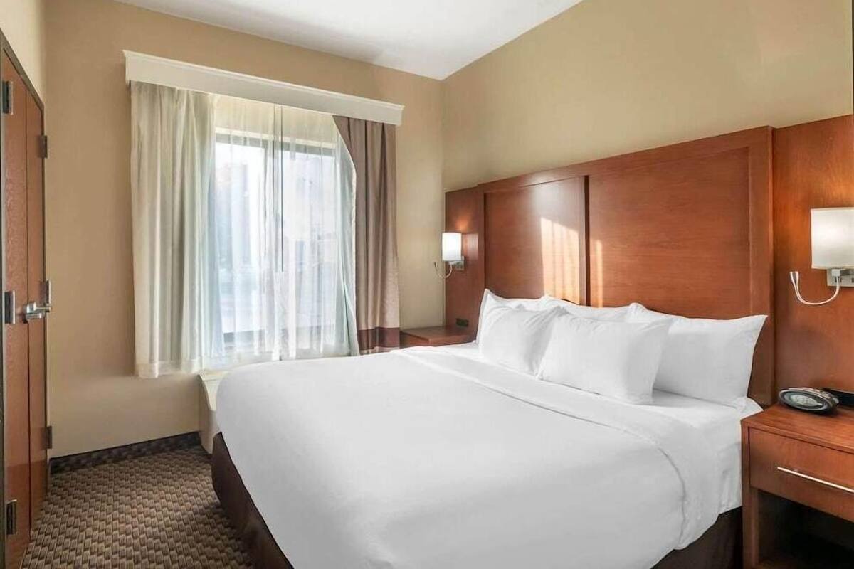 A hotel room with a large bed, a wooden headboard, a bedside table with a lamp, and a window with curtains.