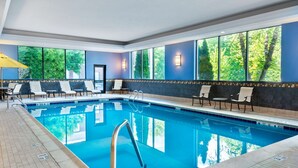 Ein Hallenbad mit klarem, blauem Wasser, umgeben von Liegestühlen und großen Fenstern mit Blick auf die grüne Umgebung.