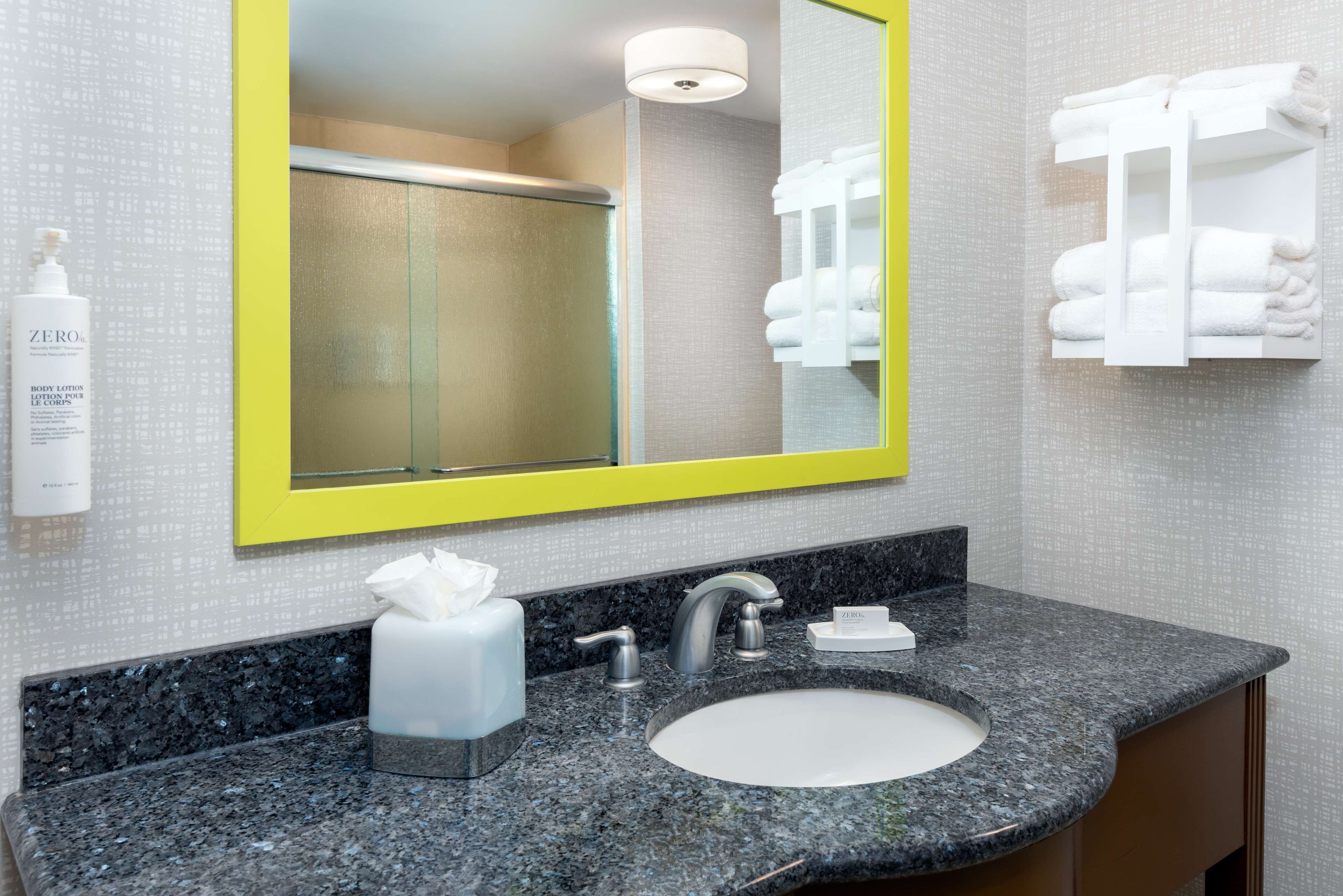 Un baño con encimera de granito, un lavabo, un espejo con marco amarillo, un perchero y una botella de loción corporal Zero.