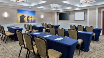A conference room with tables and chairs, a painting of a bridge, a TV, and a whiteboard.