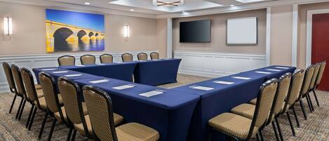 A conference room with a long table, chairs, a large painting, a TV, and a patterned carpet.