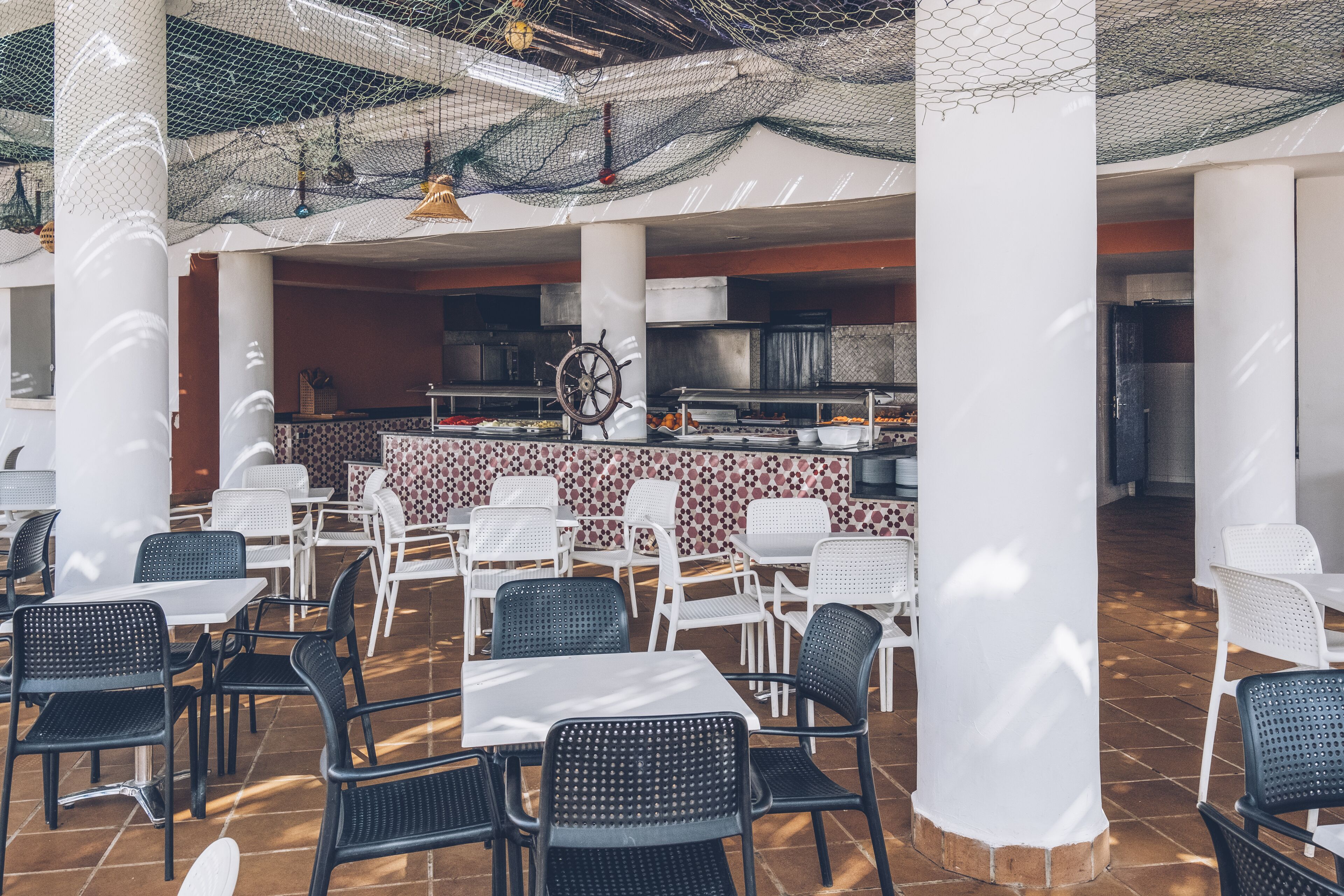 An outdoor dining area with white and black chairs, a central bar with a ship's wheel, and a tiled floor.