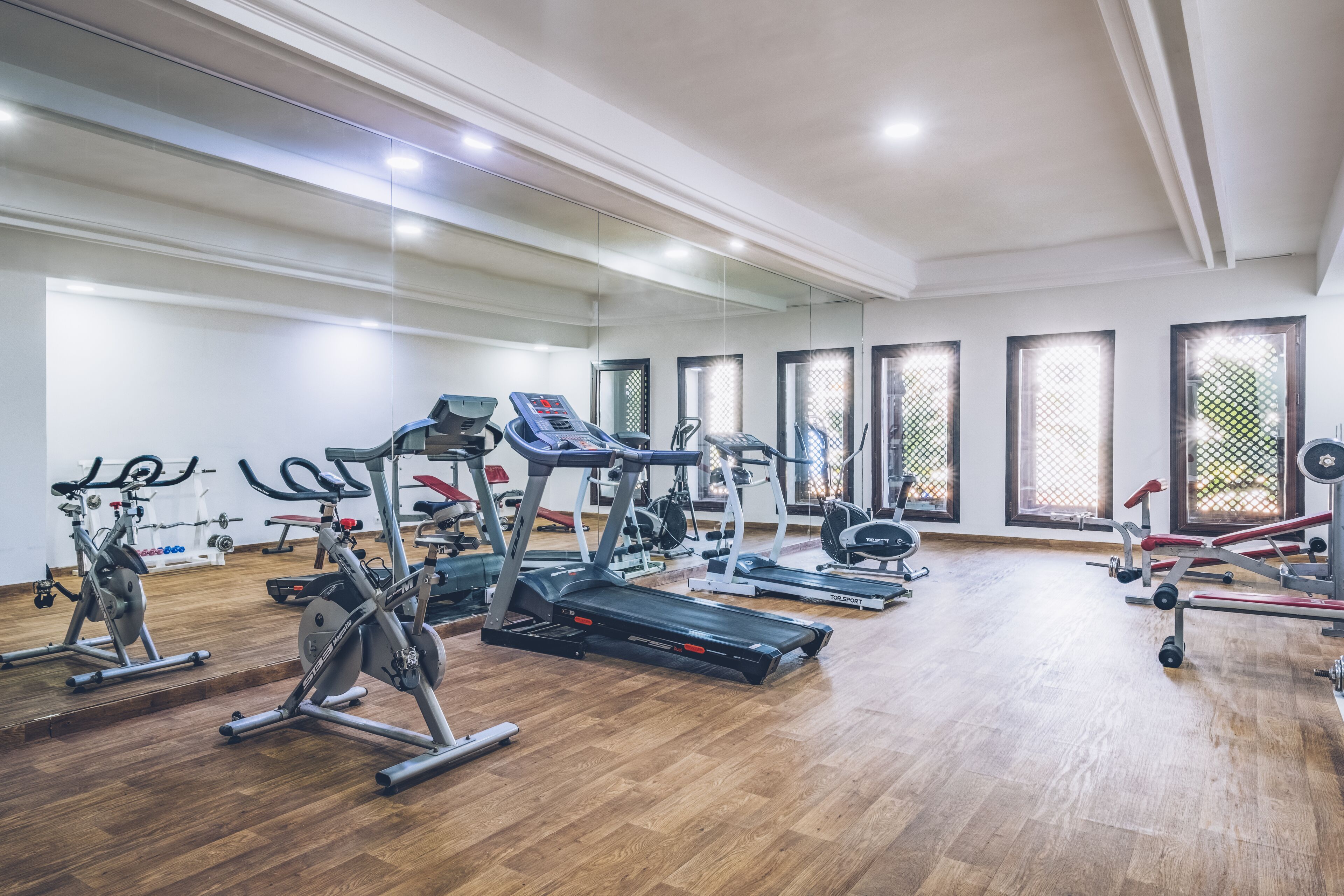 Een sportschool met diverse fitnessapparaten, grote ramen en een houten vloer.