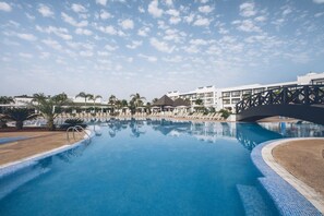 A large swimming pool with a bridge, surrounded by palm trees and a building with balconies. - Iberostar Waves Saïdia (Saidia)