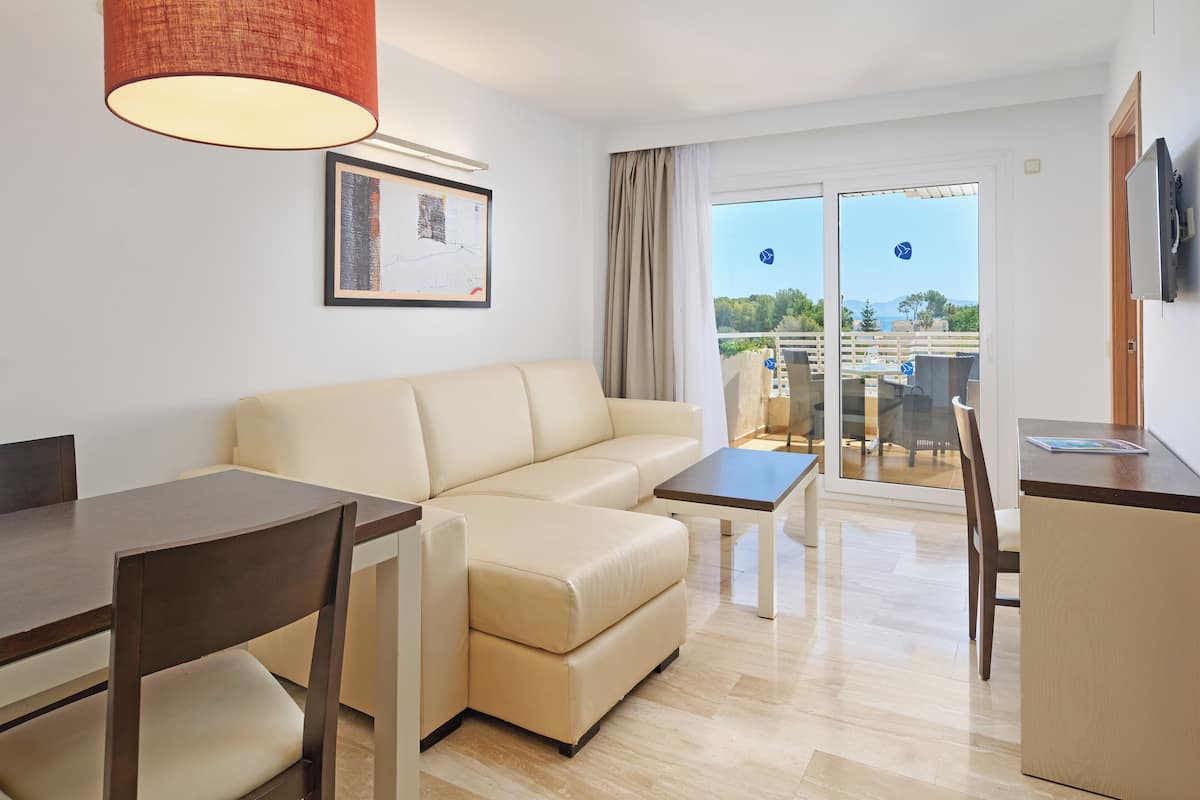 A modern living room with a sofa, dining table, and chairs. There is a TV mounted on the wall and a window with curtains.
