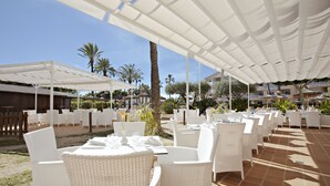 An outdoor dining area with white wicker tables and chairs, covered by white canopies, set against a backdrop of palm trees and buildings. - Grupotel Port d'Alcudia (Alcúdia)