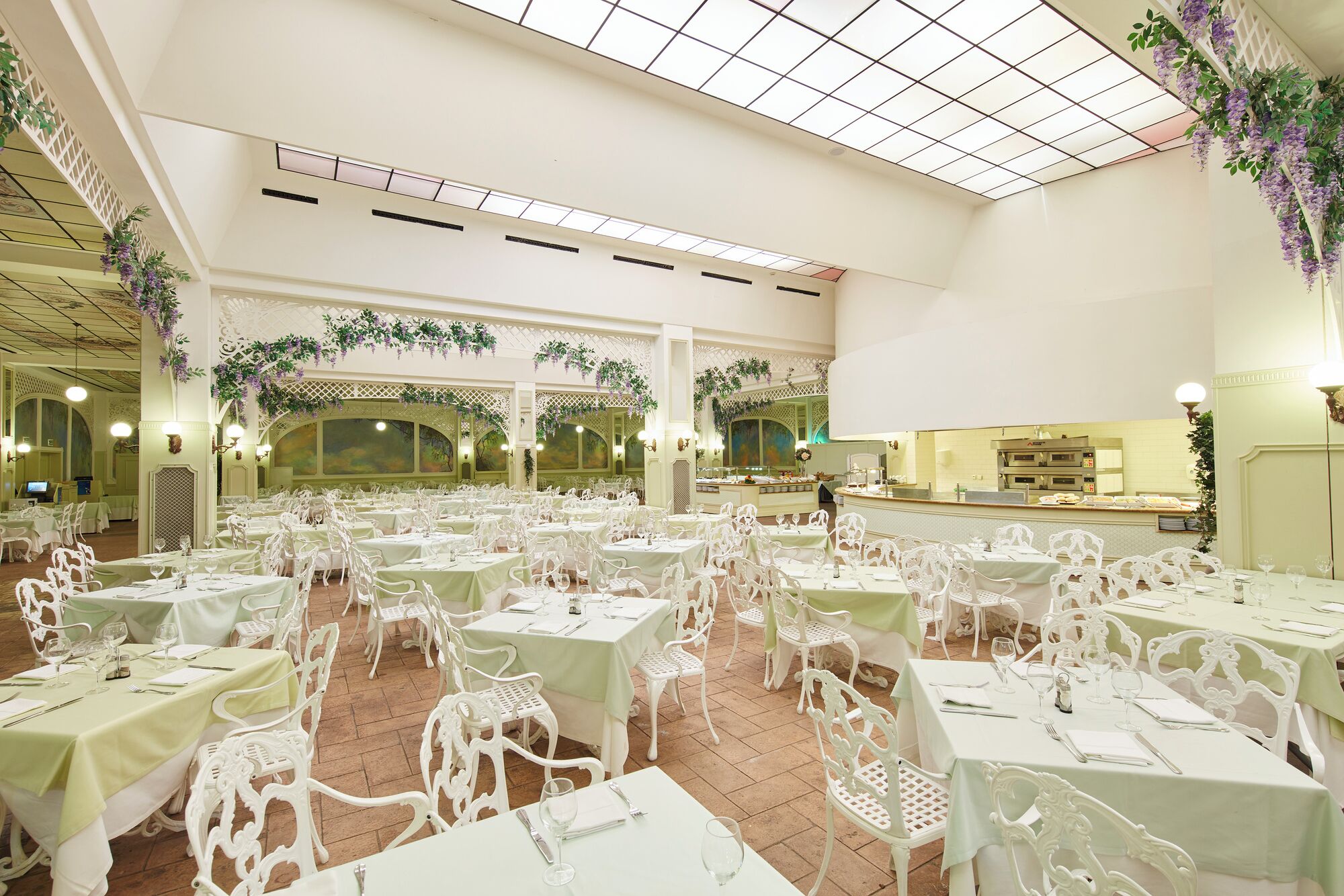 Un amplio salón de banquetes con varias mesas y sillas, un techo alto con tragaluces y decoración con plantas.