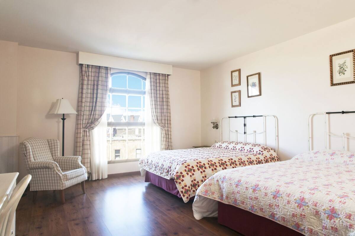 Un dormitorio con dos camas, una silla, una lámpara y una ventana con cortinas.