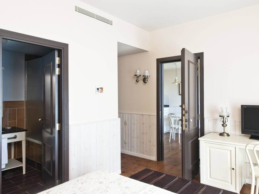 Habitación de hotel con una puerta de madera oscura, un mueble blanco con televisor de pantalla plana, una silla blanca y una pequeña mesa.