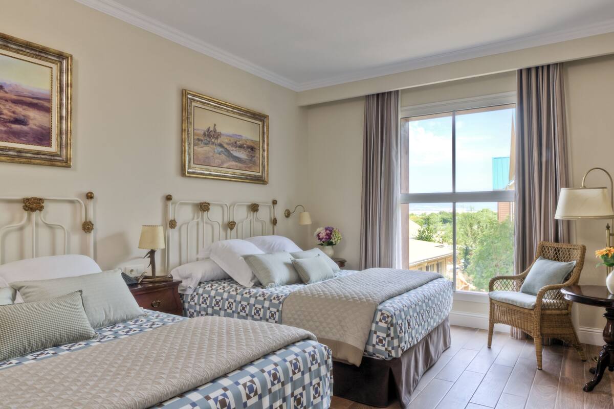Una habitación de hotel con dos camas, un ventanal con cortinas, una silla de mimbre y una mesita con lámpara.