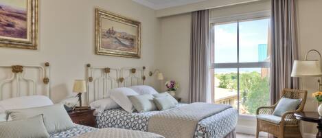 A hotel room with two beds, a large window with curtains, a wicker chair, and a small table with a lamp.