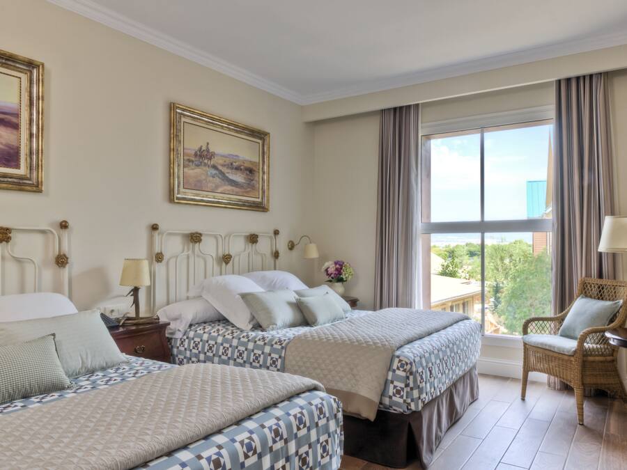 A hotel room with two beds, a large window with curtains, a wicker chair, and a small table with a lamp.