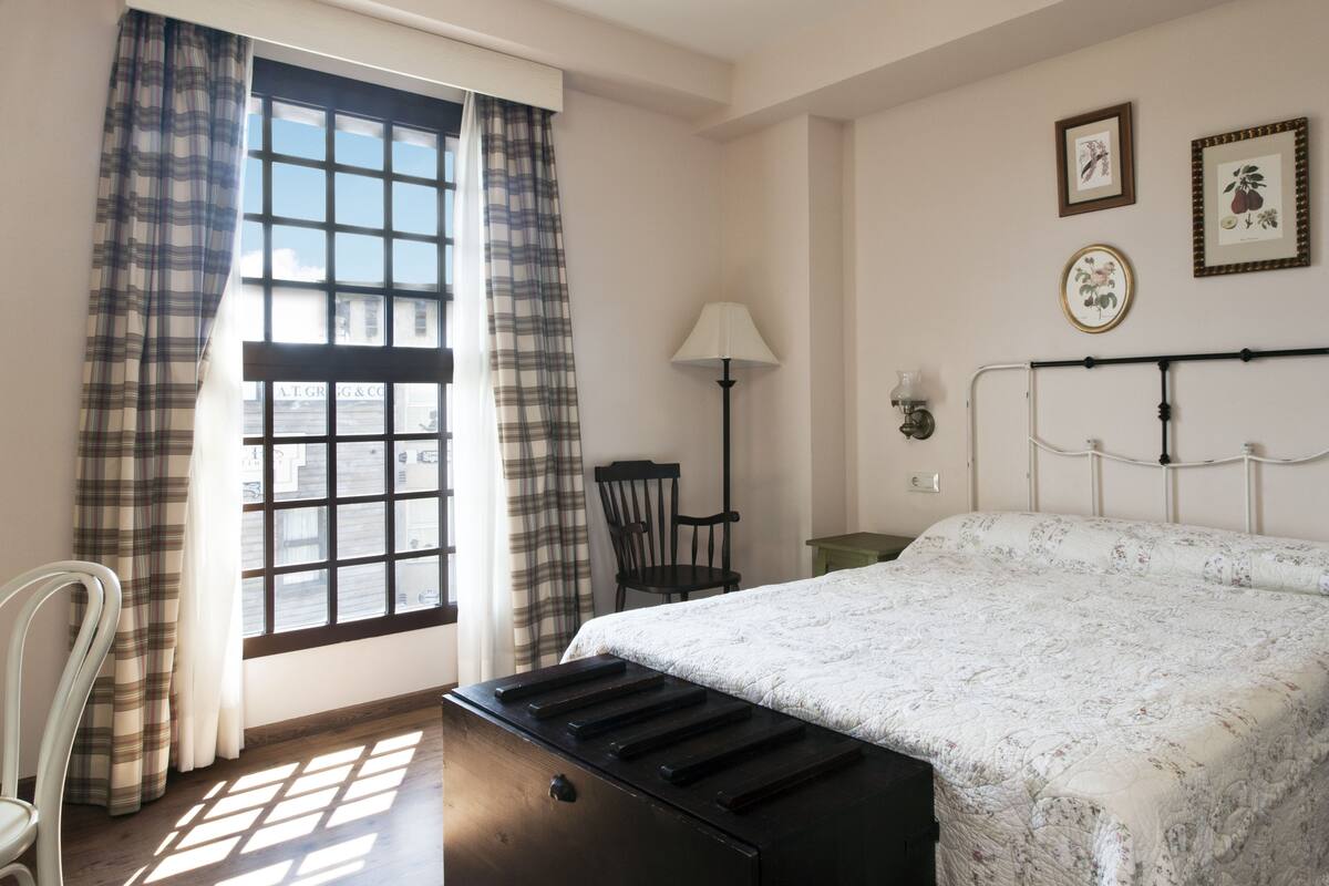 Un dormitorio con cama, una silla, un escritorio, una lámpara y una ventana con cortinas.