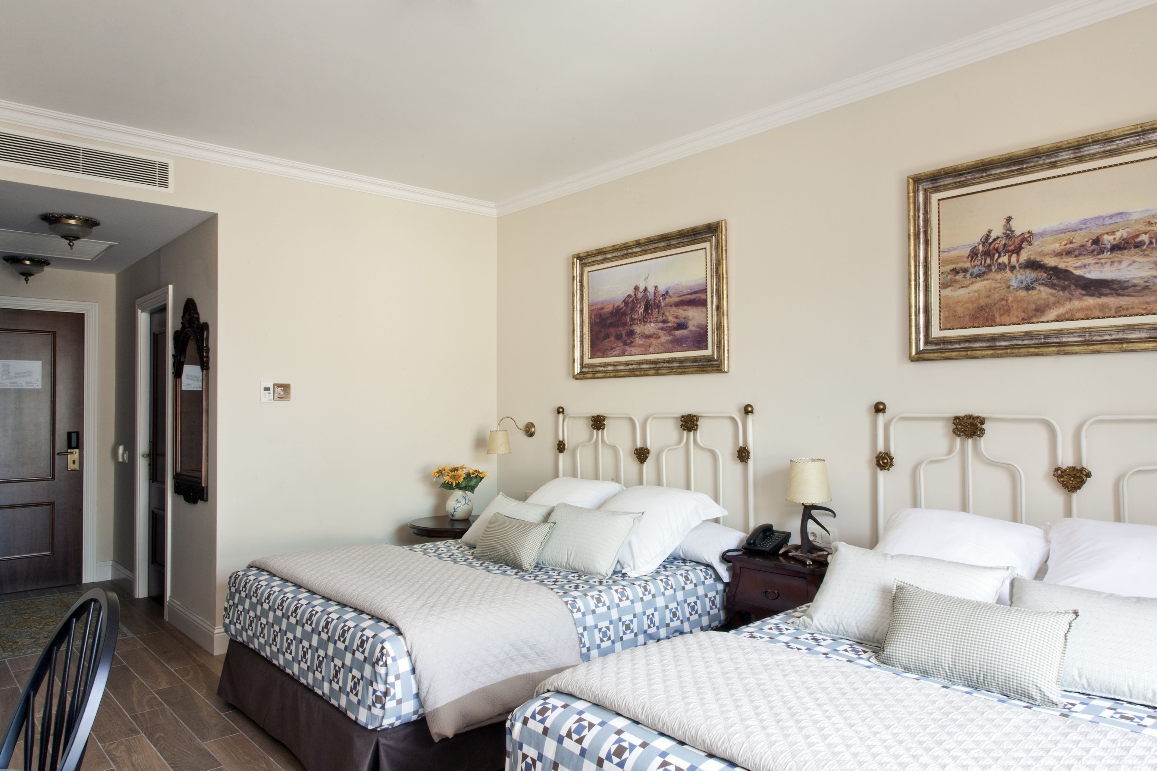 Una habitación de hotel con dos camas, una puerta de madera, una mesita de noche con lámpara y dos cuadros enmarcados en la pared.